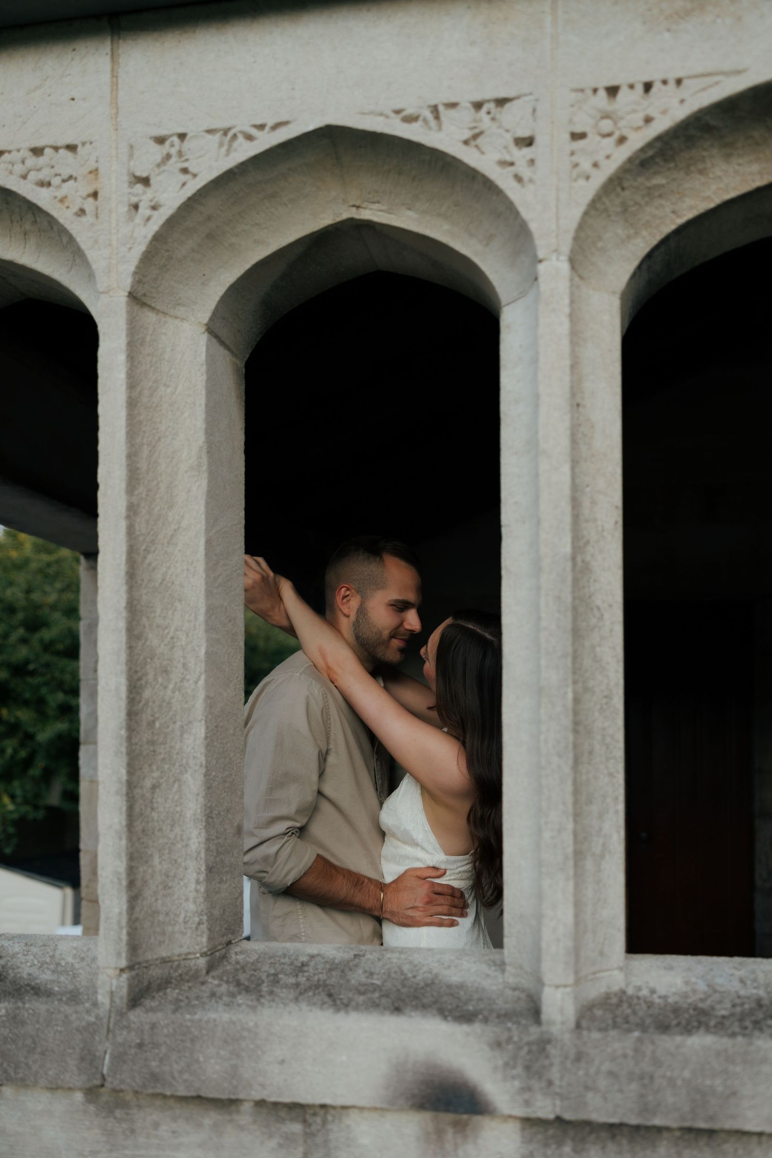 A romantic couple embraces and nearly kisses framed by elegant stone arched columns at dusk.