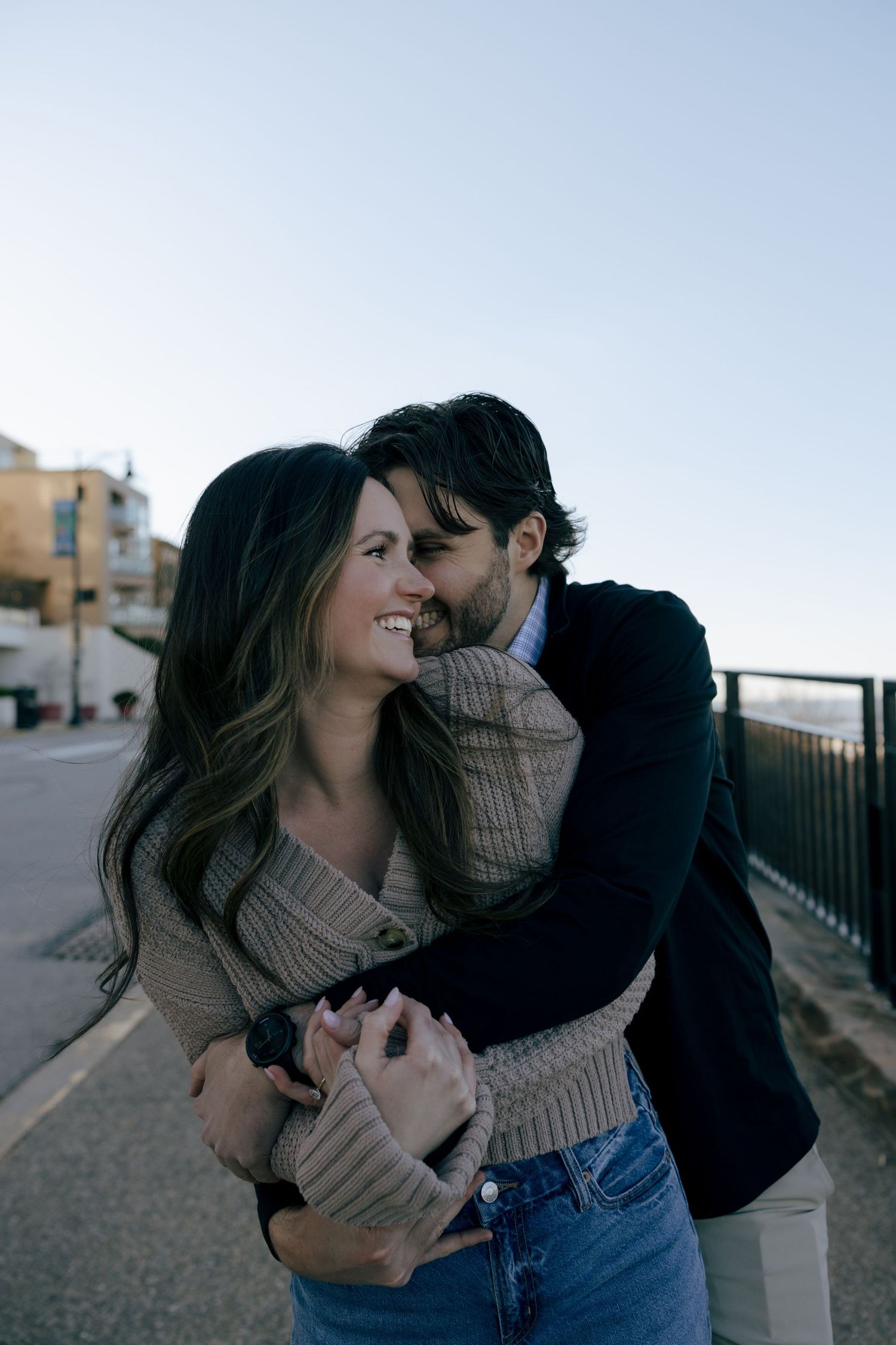 A couple embraces and laughs together on an urban street while wearing cozy sweaters at dusk.