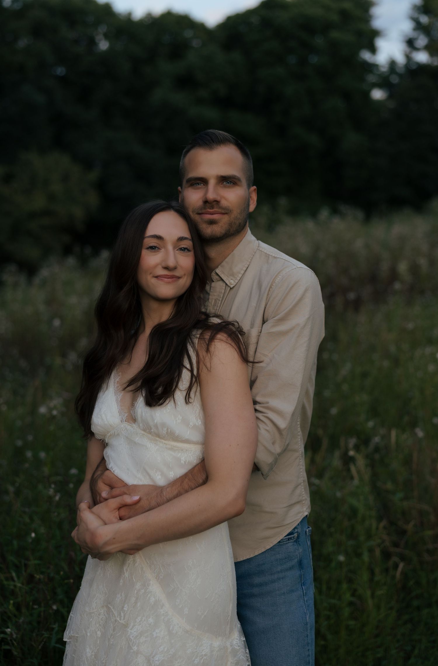 Romantic couple posing in a wildflower field at twilight, woman in white dress held from behind by man.
