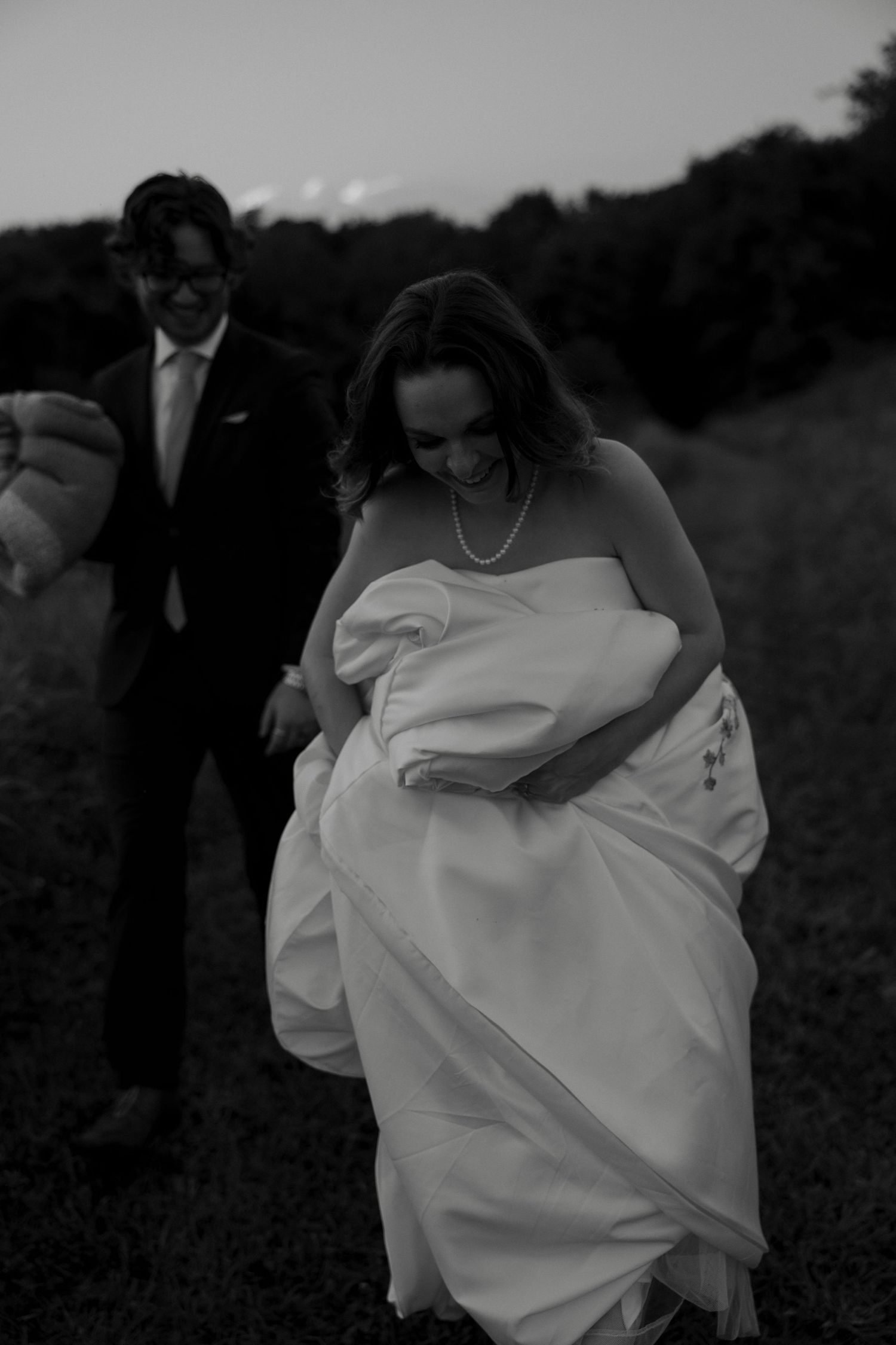 Black and white photo of joyful bride and groom walking together along an outdoor path at twilight.