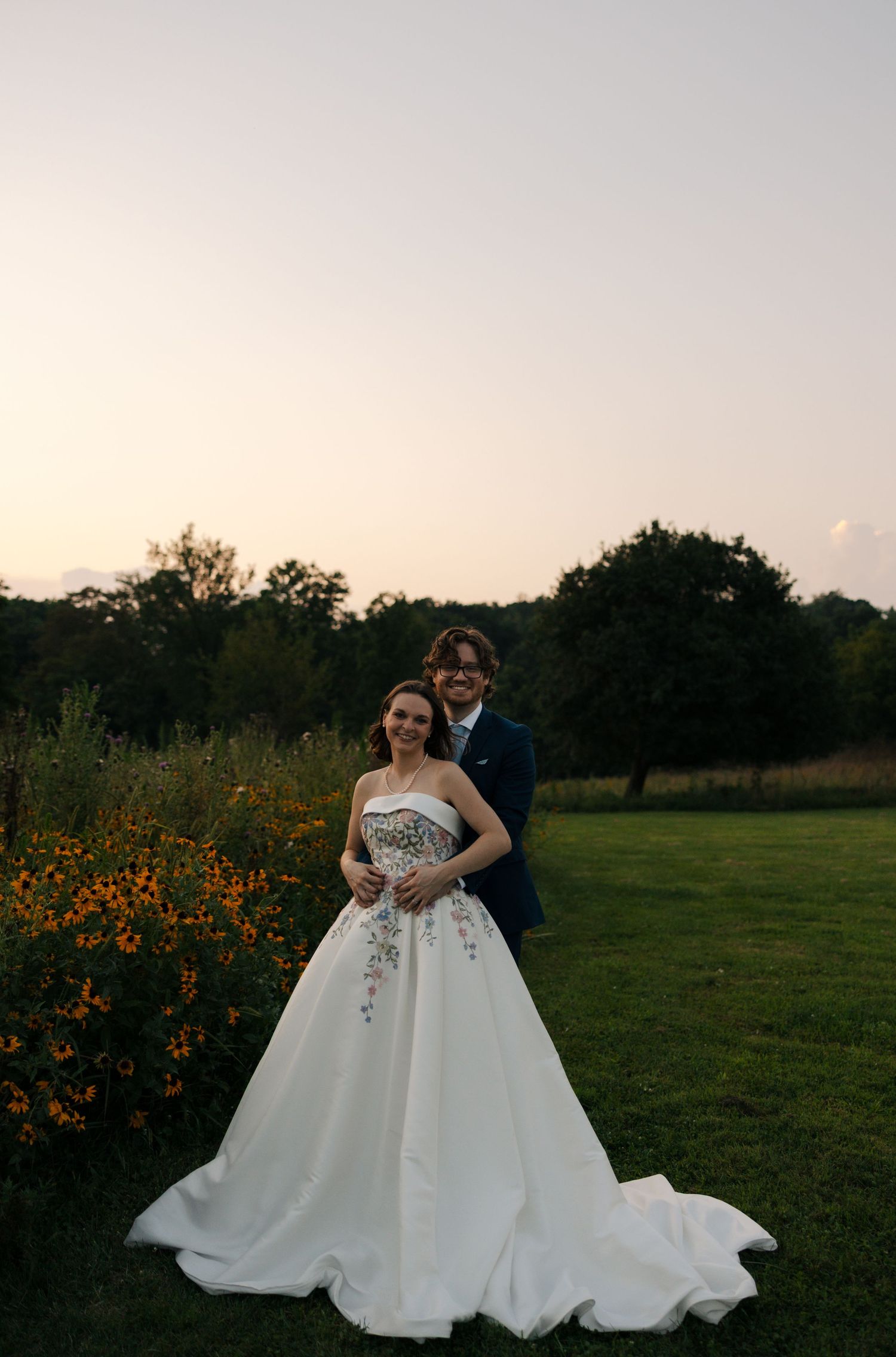 Bride and groom pose together near orange wildflowers at sunset, she in a floral gown, he in navy suit.