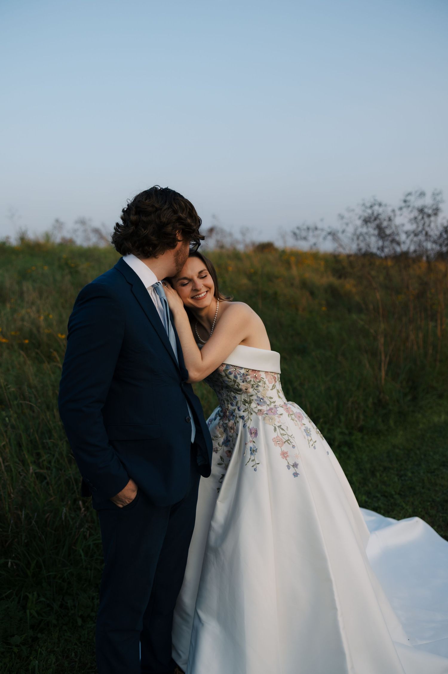 Groom kisses brides cheek as she smiles leaning against him in a sunlit meadow.