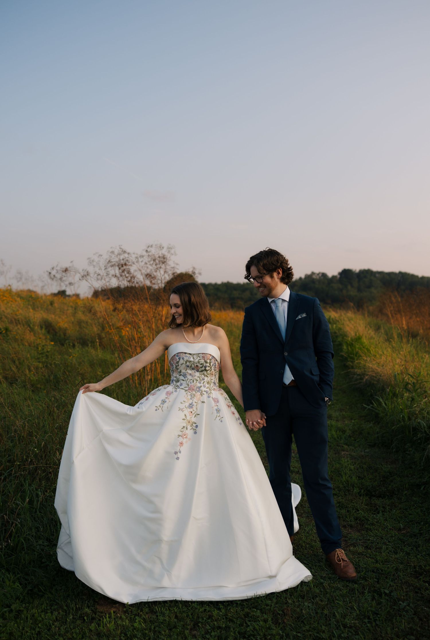 Bride in white floral gown holds dress while walking hand-in-hand with groom in navy suit through green meadow.