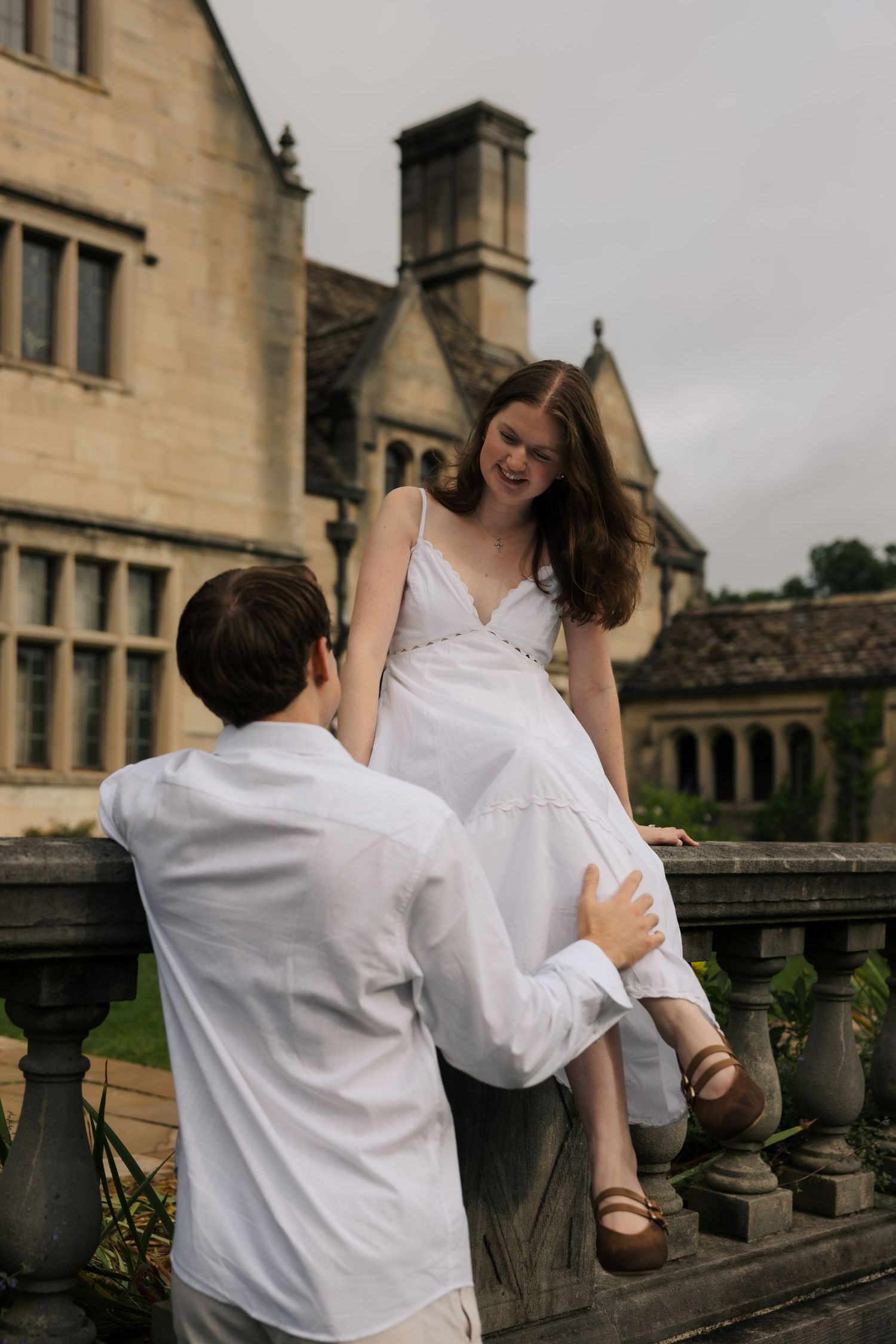Young woman in white dress sitting on stone balustrade while man in white shirt reaches up to her.