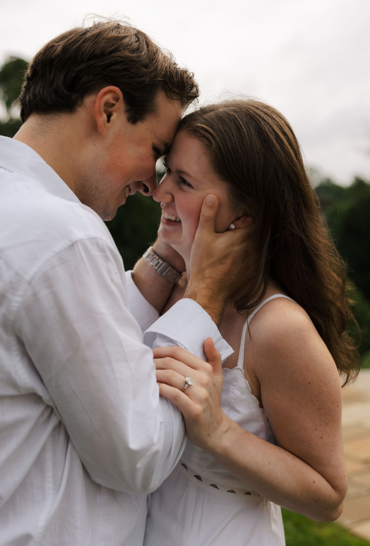 Happy couple in white outfits embrace forehead-to-forehead, laughing intimately outdoors.