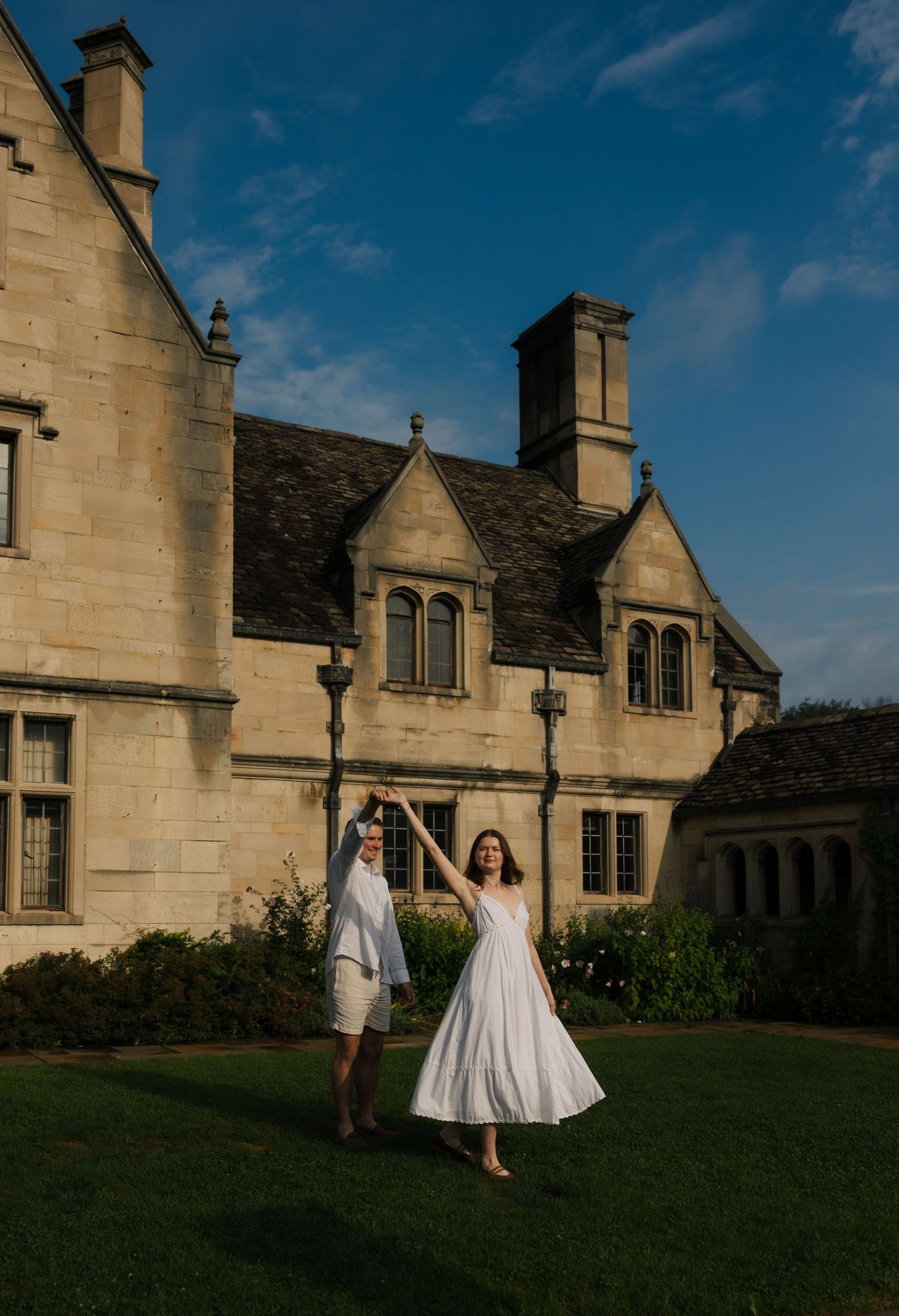 Two people in white outfits hold hands on a lawn before a grand English manor under a blue sky.