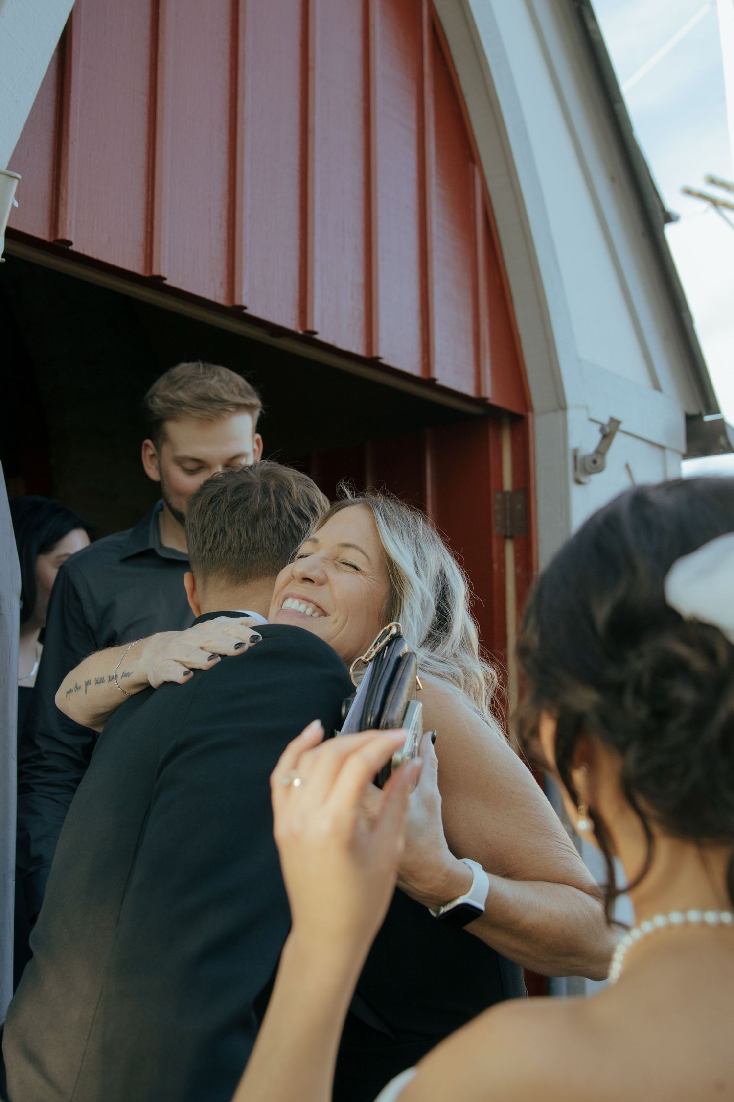 Joyful wedding guest embracing the groom at the entrance of a rustic red barn venue.