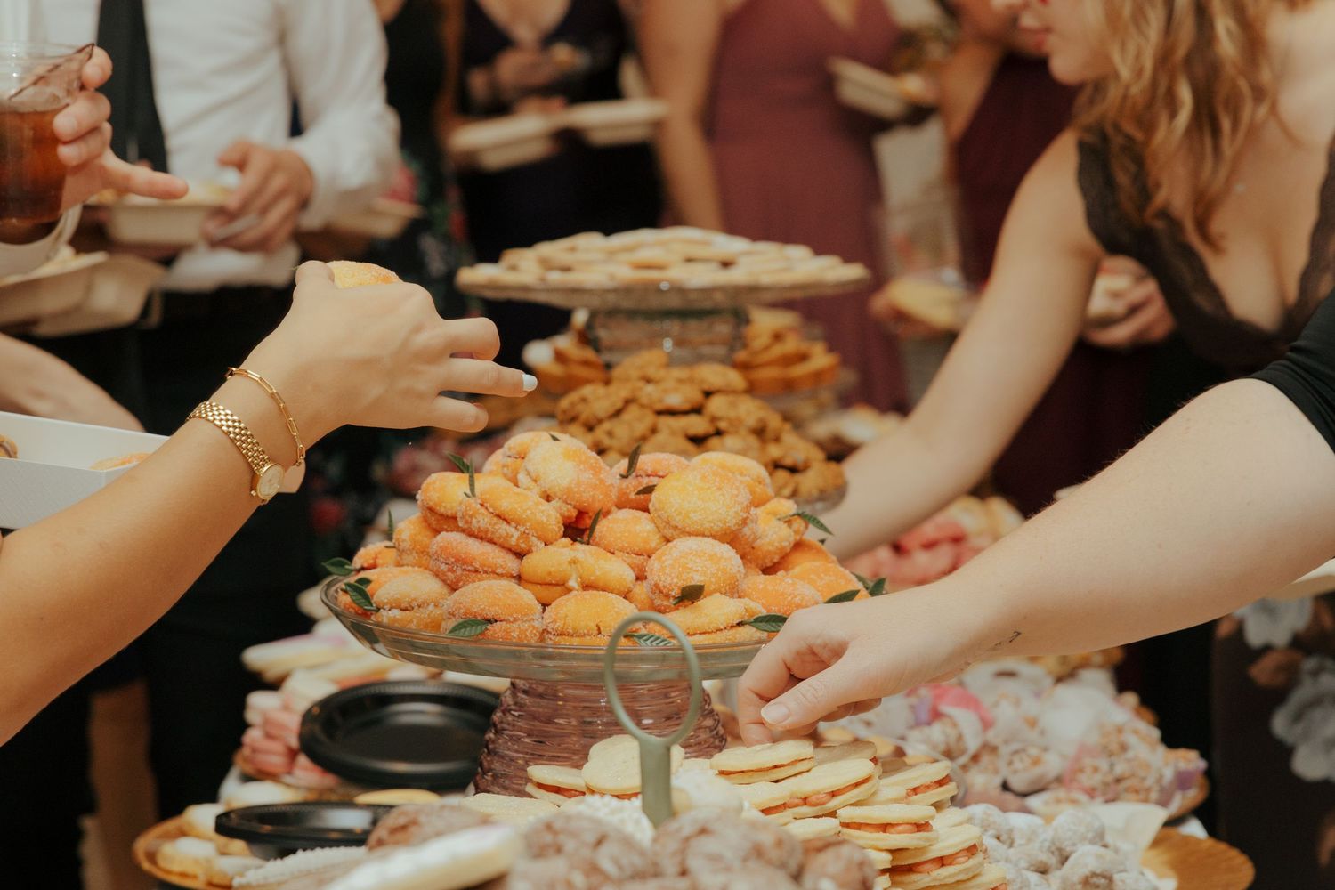 Wedding guests reaching for pastries and sweets at beautifully arranged dessert buffet table during reception.