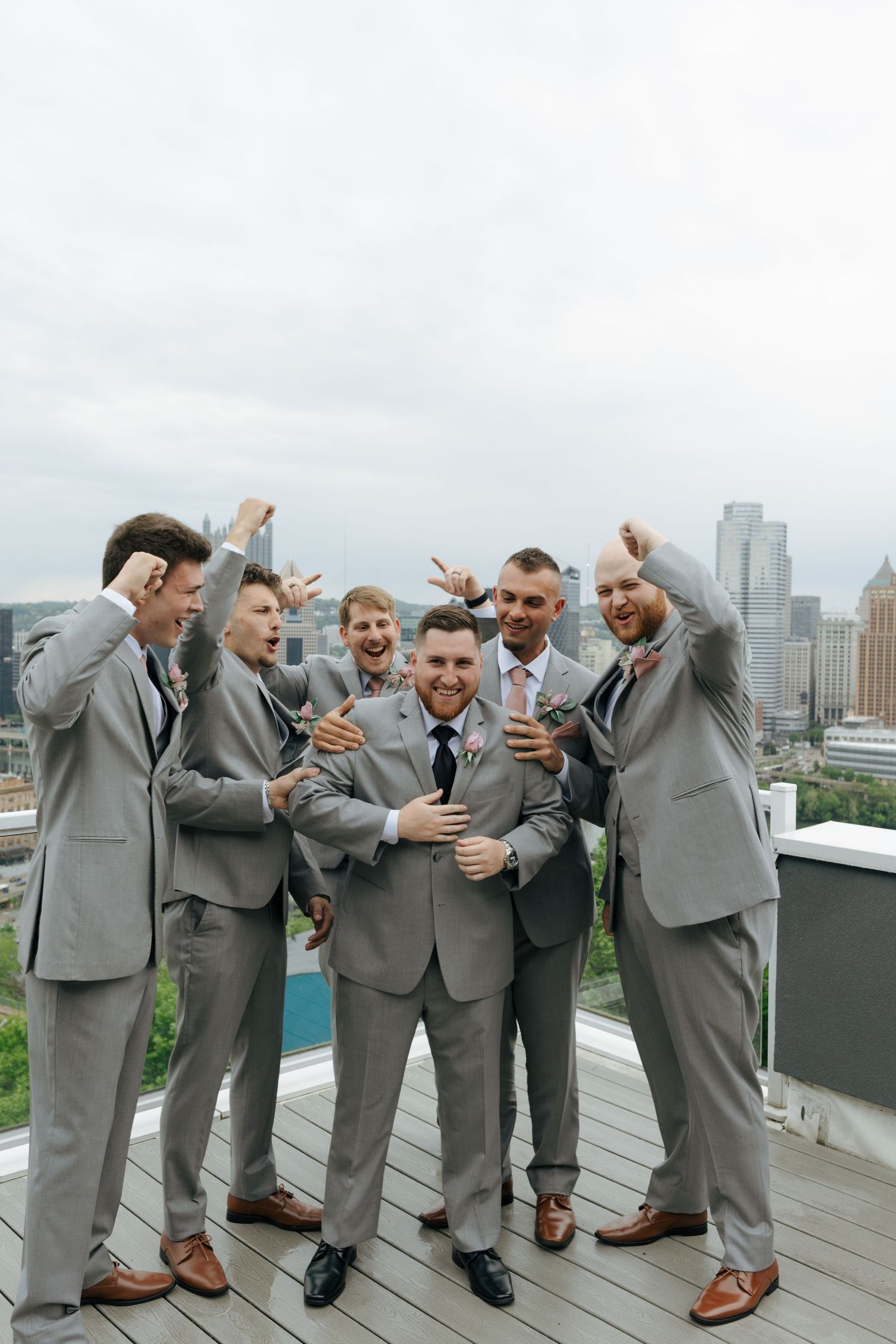 Groom and groomsmen in gray suits celebrate on a rooftop with a city skyline backdrop.