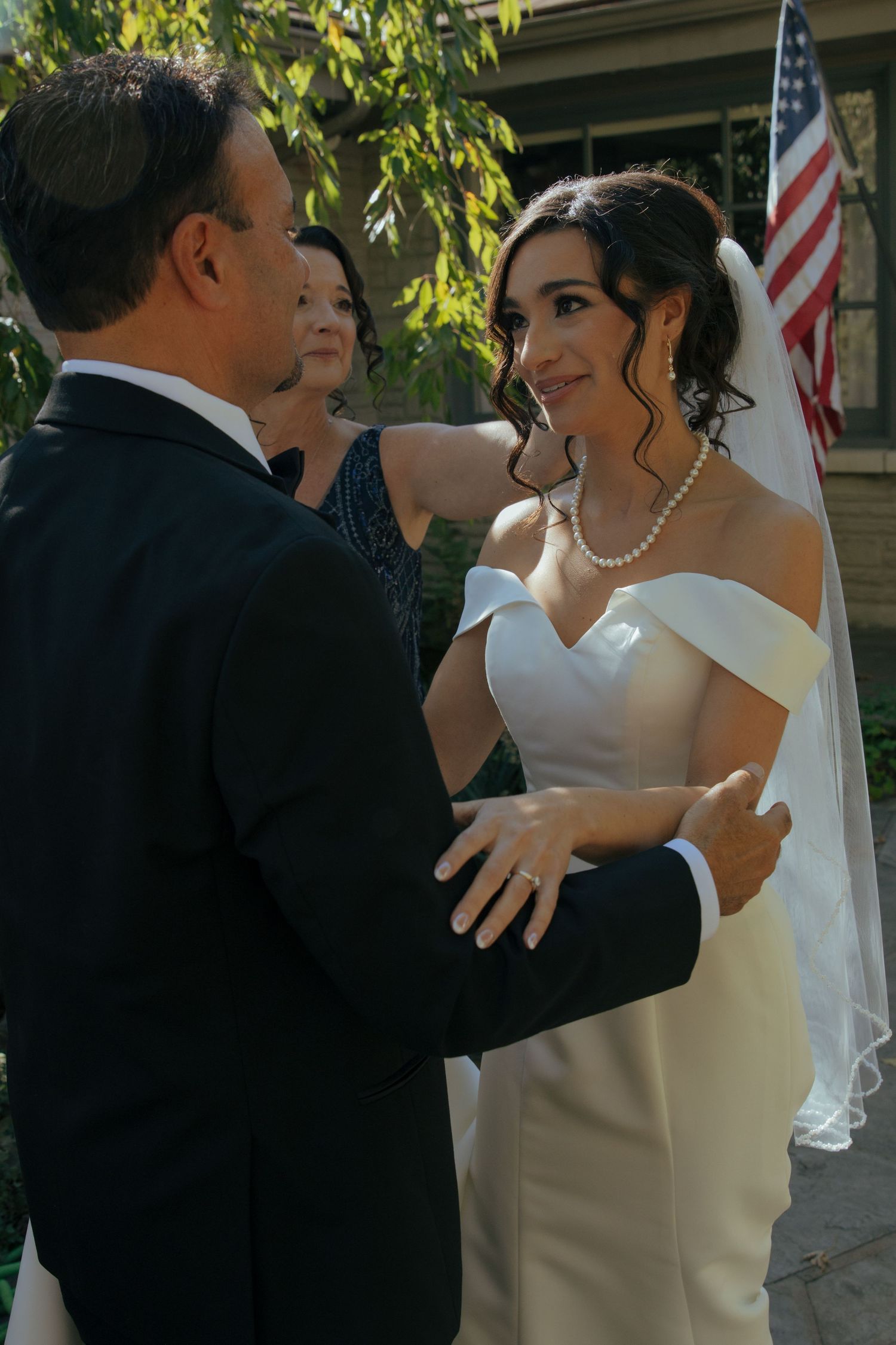 Bride in off-shoulder white gown and veil smiles at groom in tuxedo during outdoor wedding ceremony.