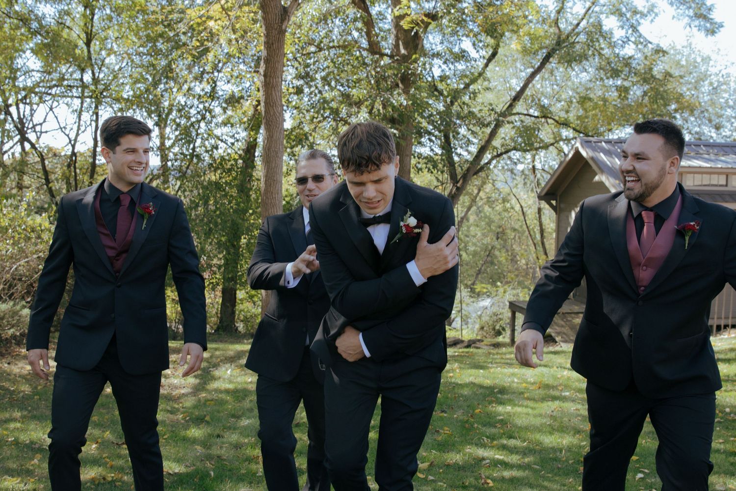 Four groomsmen in black suits with burgundy ties laughing and celebrating outdoors at a wedding.