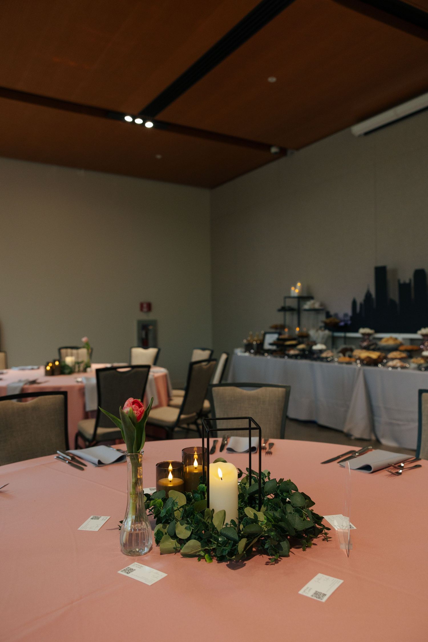 Elegant wedding reception table with pink linen, candle centerpiece, greenery wreath, and floral arrangement in softly lit venue.