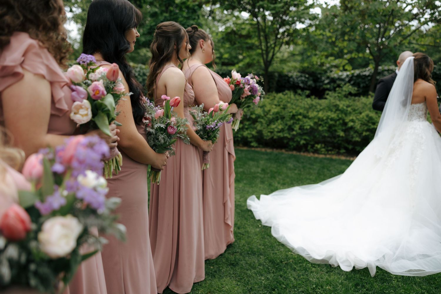 Bridesmaids in dusty rose gowns holding colorful bouquets stand beside bride in white wedding dress.