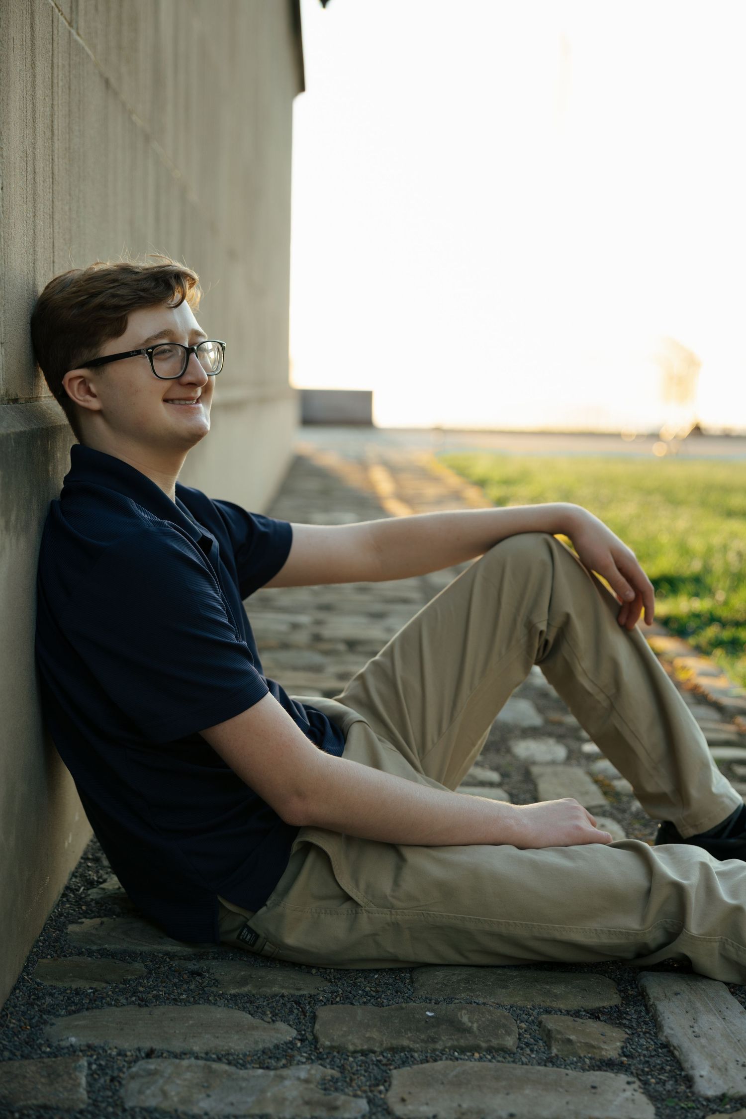 Young man in navy polo and khakis sitting against a wall on cobblestone ground at sunset.