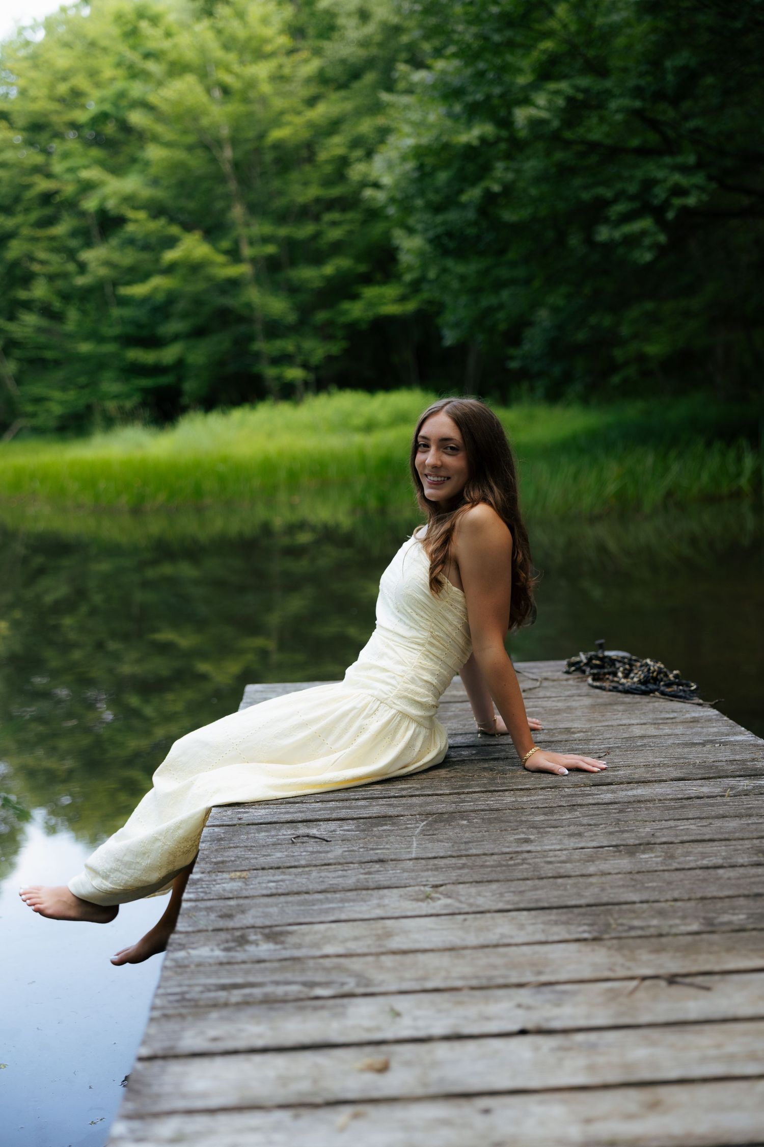 Smiling young woman in cream dress sitting on rustic wooden dock beside a serene forest lake.