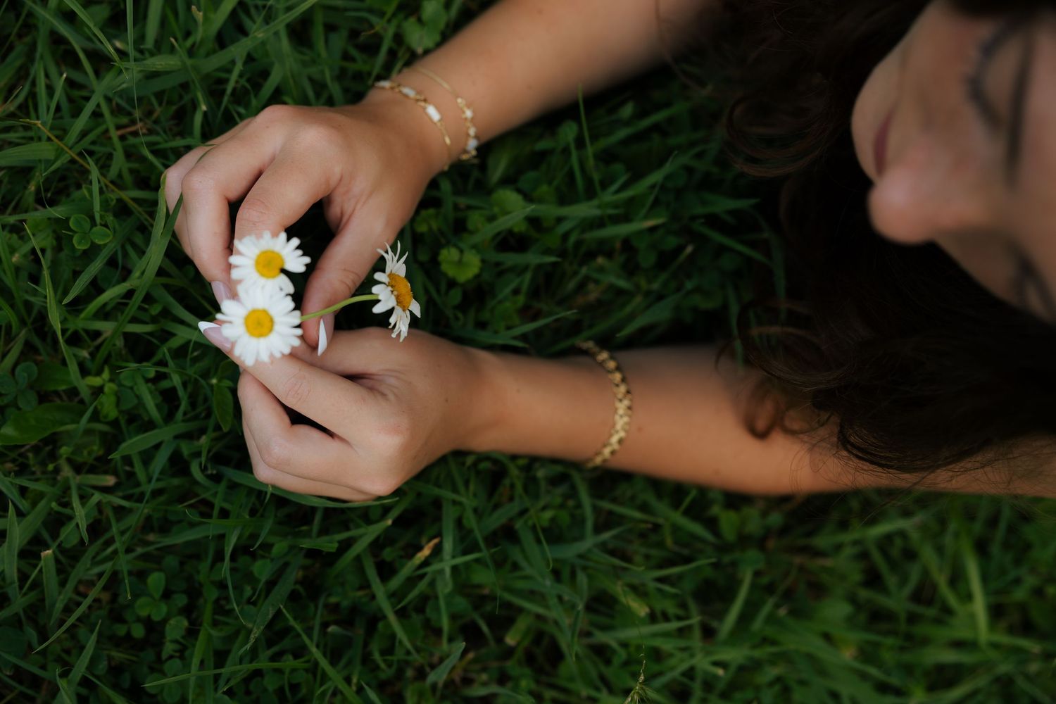 Close-up of hands with gold bracelets holding white daisy flowers over green grass.