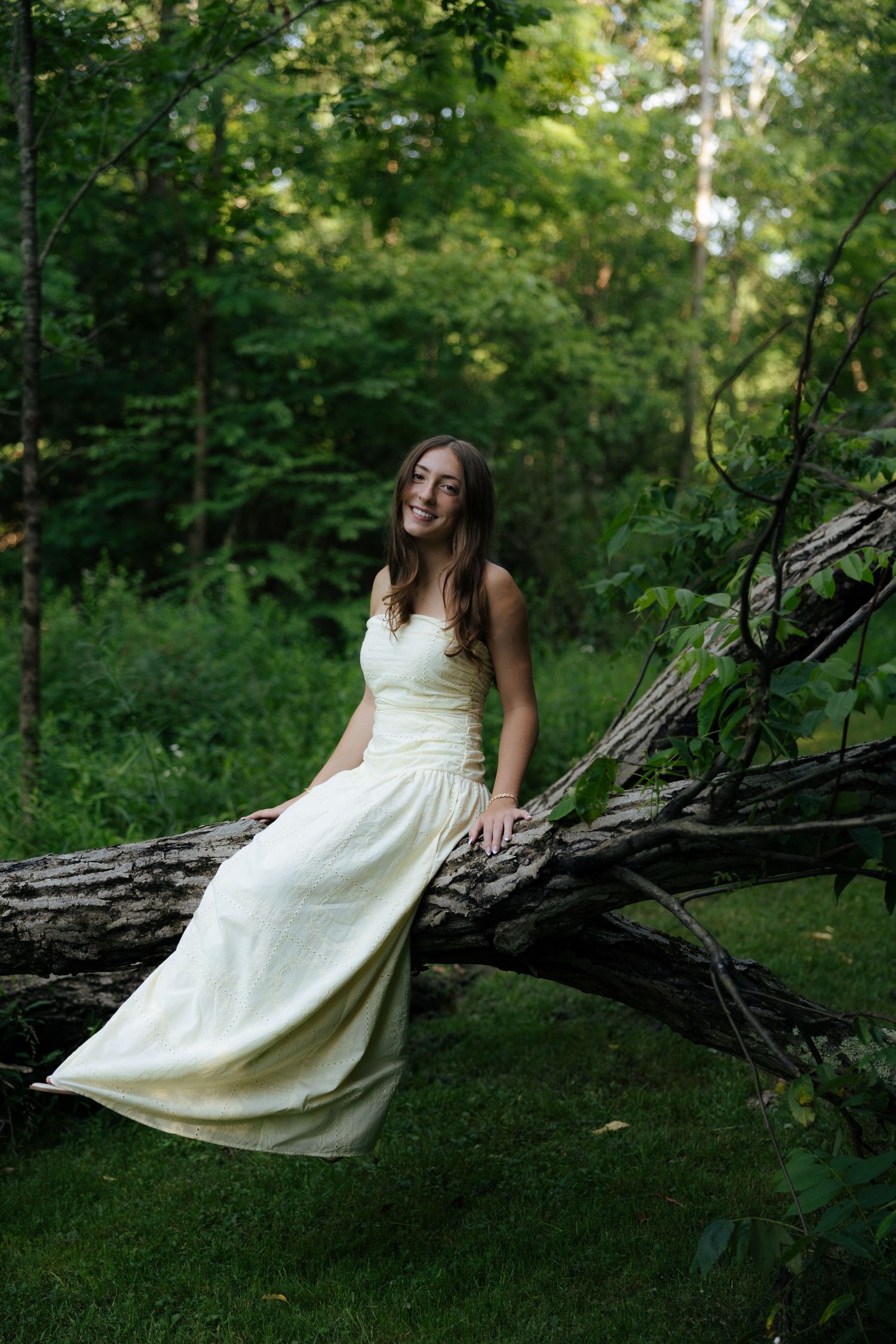 Smiling young woman in elegant white dress sitting on fallen log in green woodland setting.