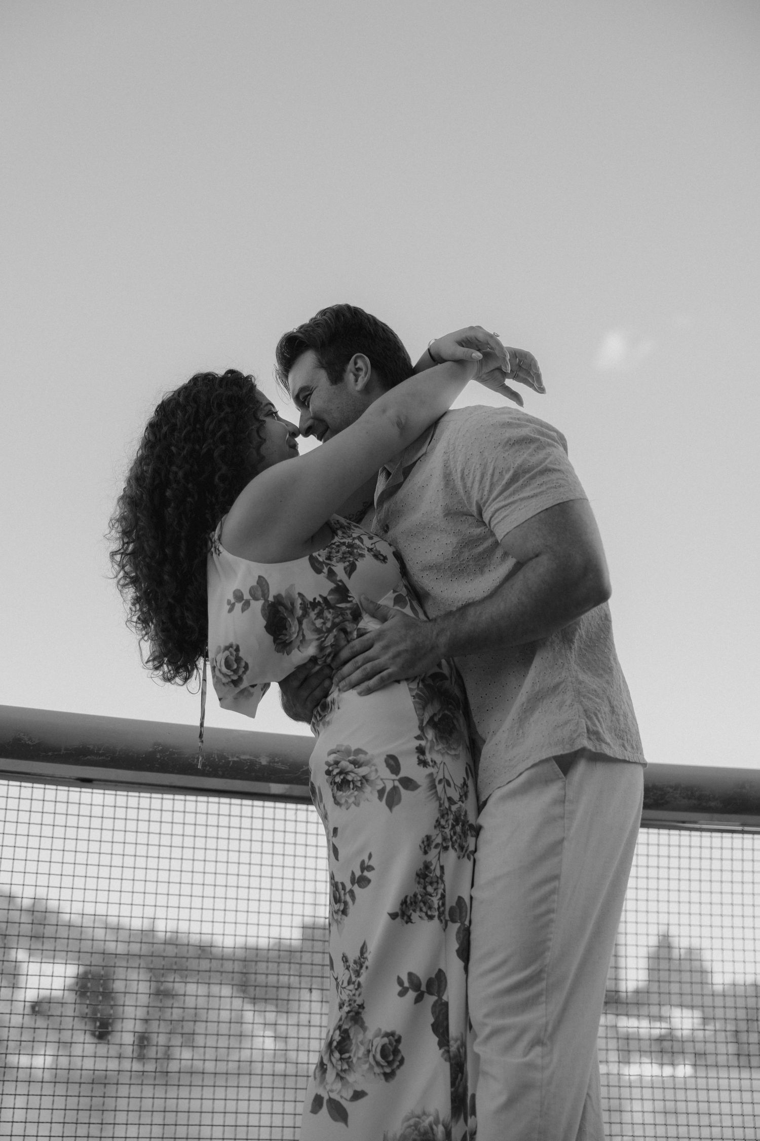 Black and white romantic photo of couple kissing on bridge railing against clear sky.