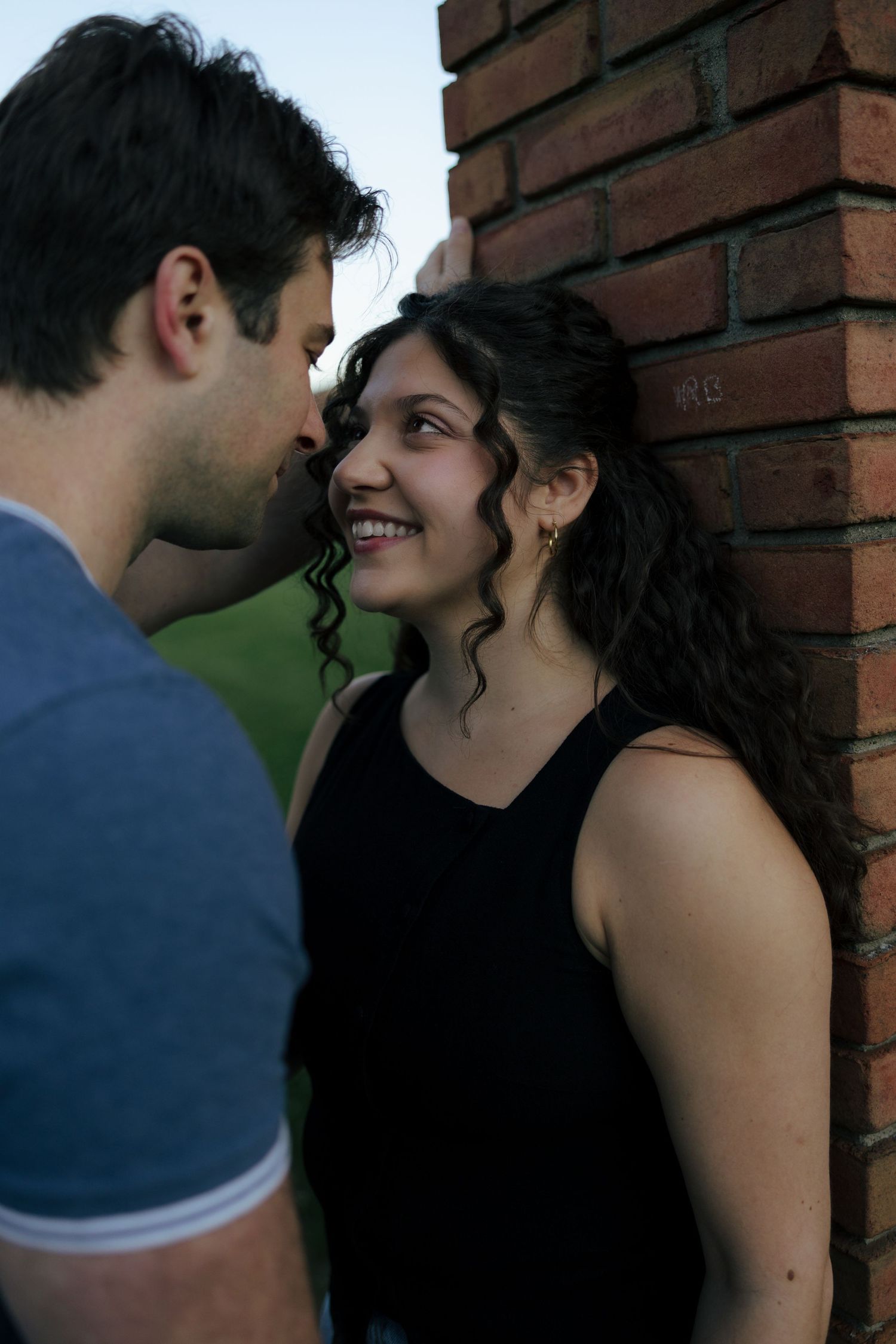 A smiling young woman leans against a brick wall as a man leans in close to her face outdoors.