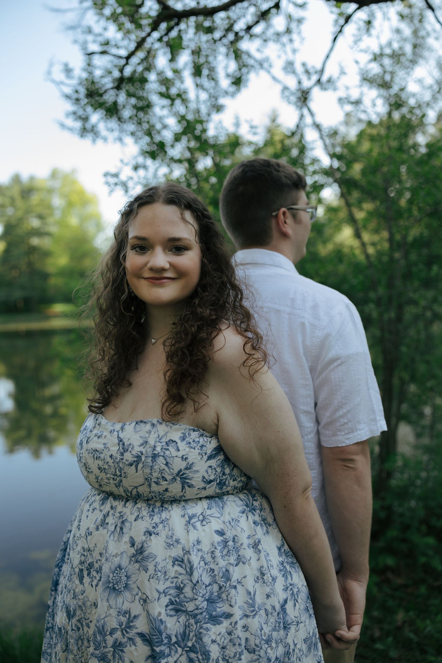 A woman in a floral dress holds hands with a man near a reflective pond surrounded by lush green trees.