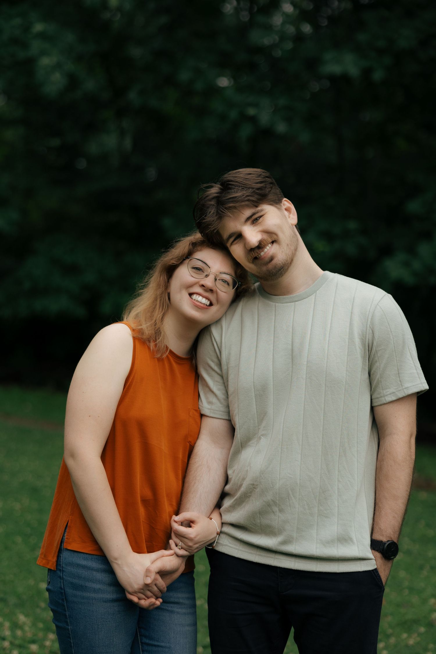 Smiling couple holds hands and leans together affectionately in a lush green outdoor park setting.