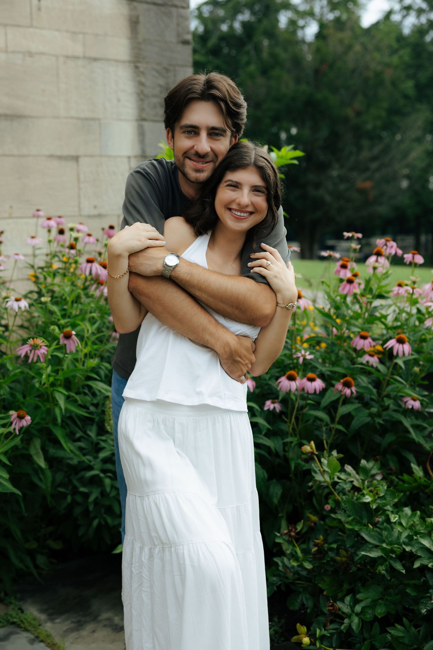 A happy couple hugs closely among vibrant pink coneflowers beside a historic stone building exterior.