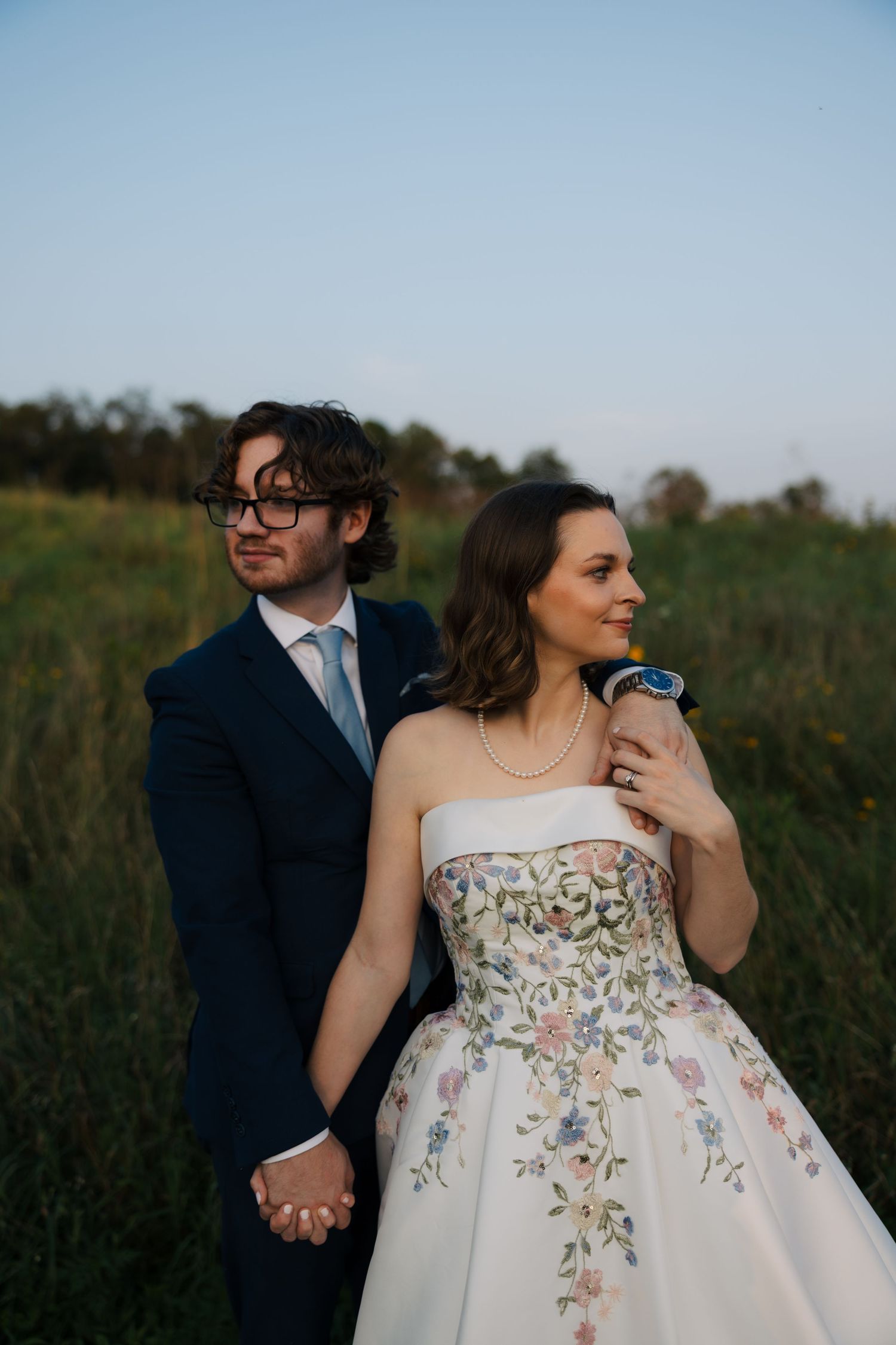 Bride and groom stand together in field at dusk, she wears floral embroidered strapless gown with pearl necklace.