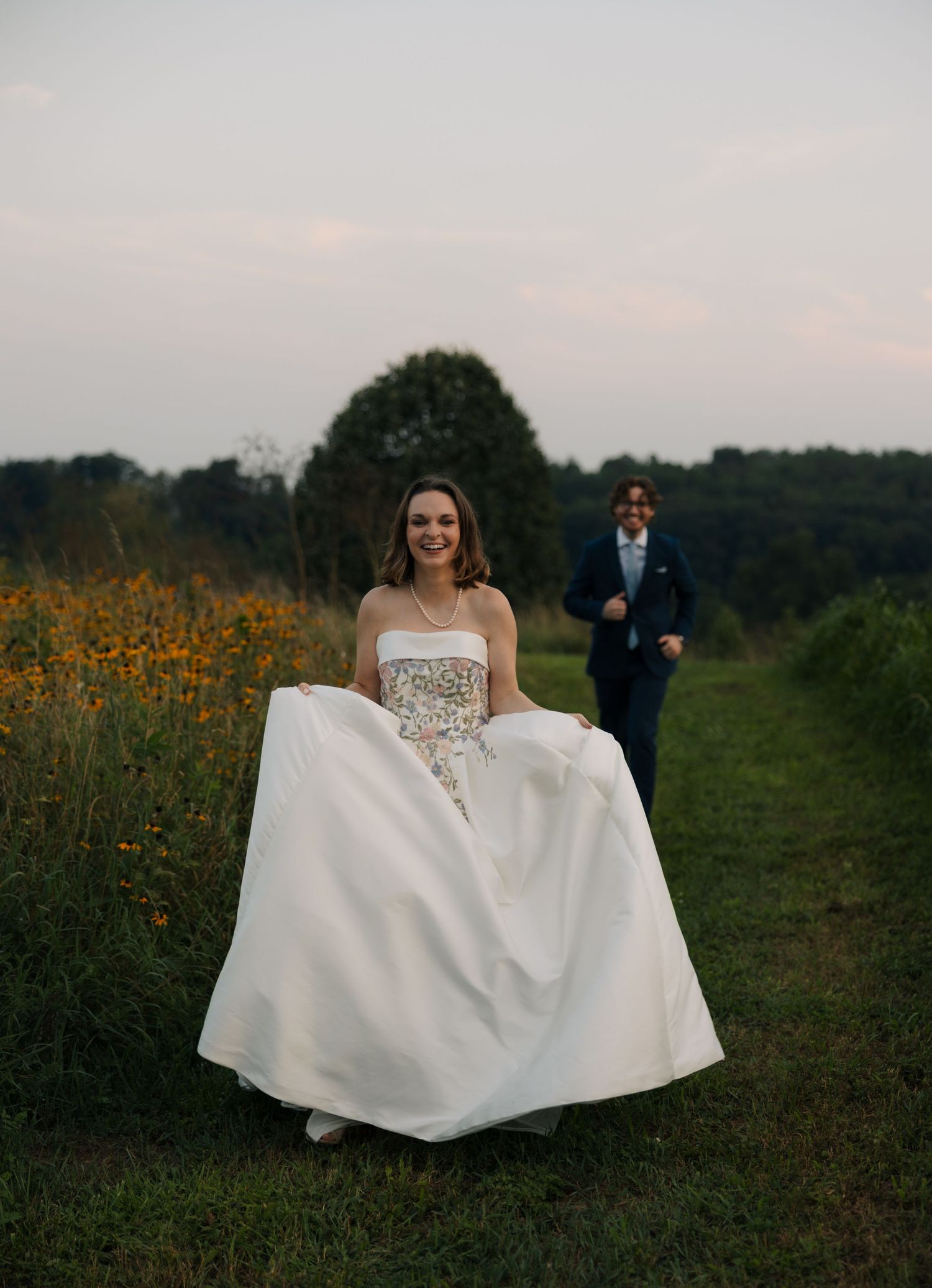 Smiling bride in white floral ballgown walks along meadow path with groom following behind at dusk.