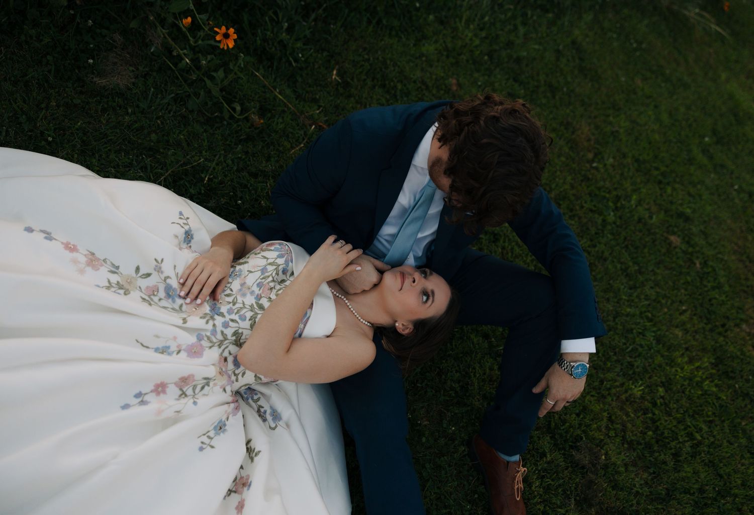 Aerial view of wedding couple lying on green grass, groom leaning to kiss bride in floral gown.