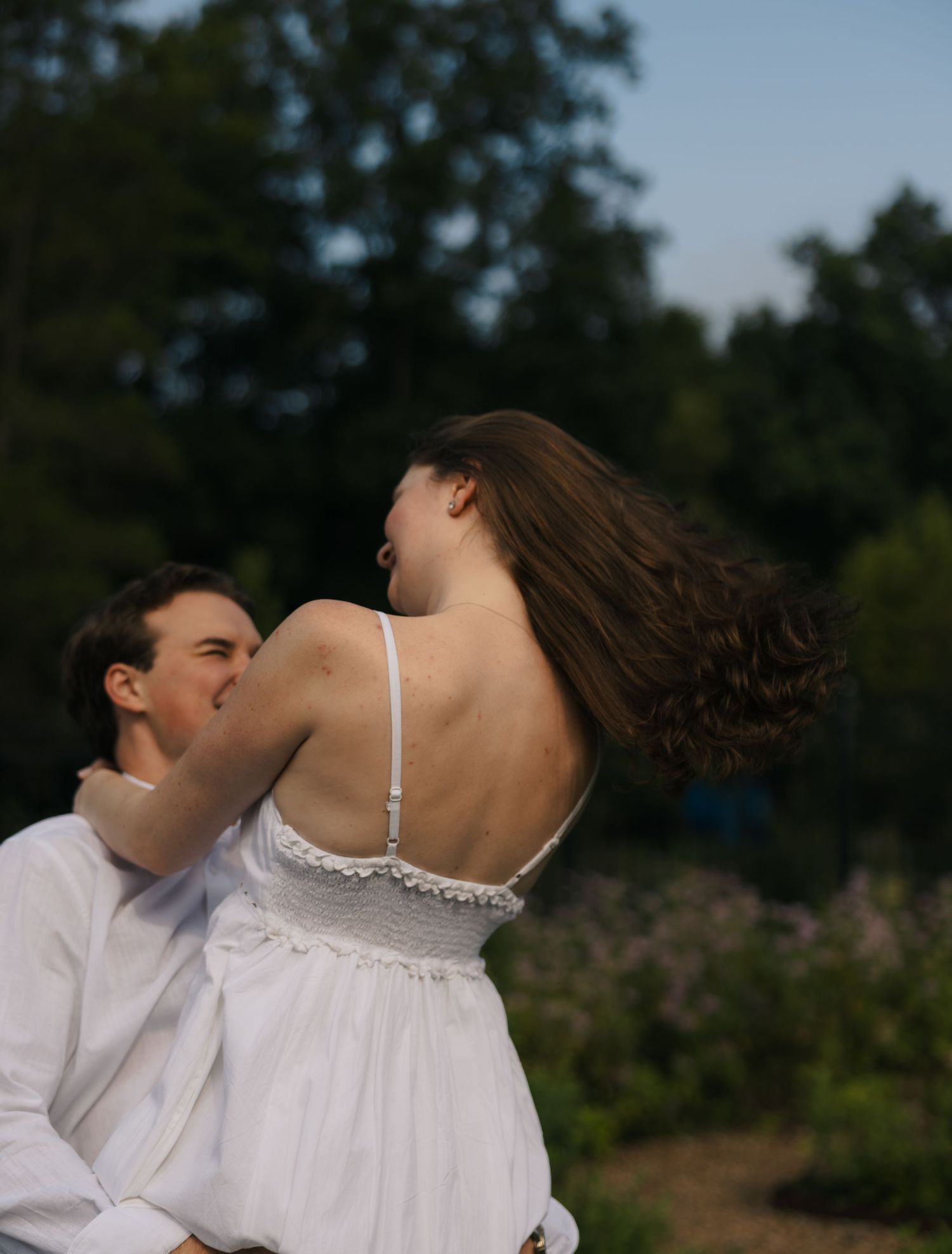 A couple in white embrace outdoors, the woman's curly hair flowing as she turns toward her partner.