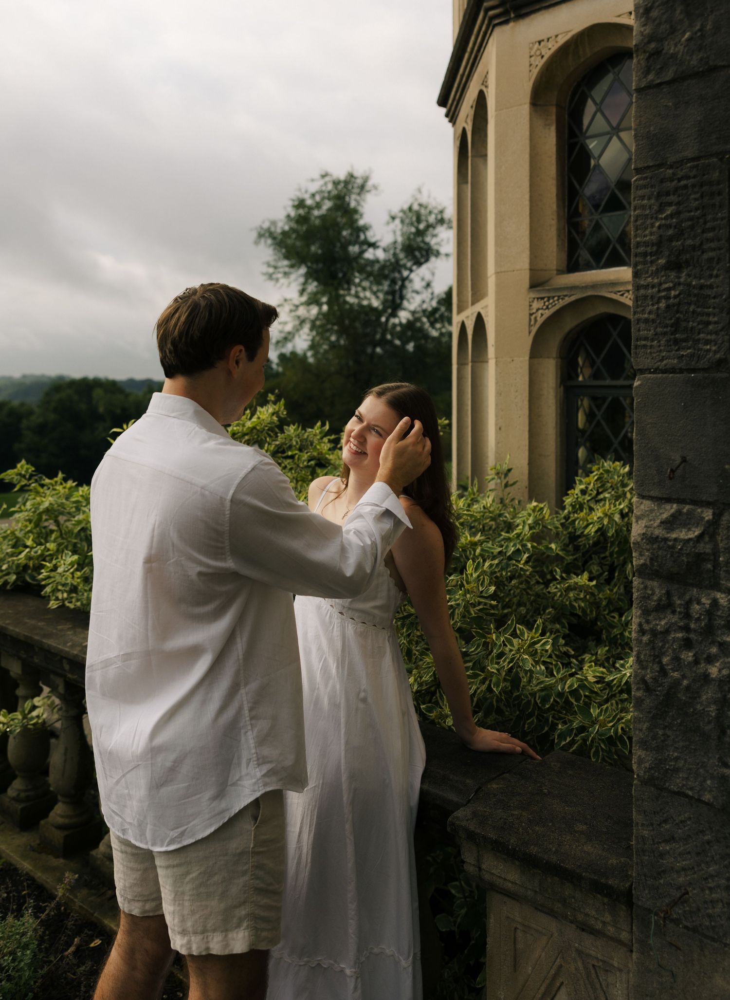 A man tenderly touches a woman's face as they share an intimate moment on a stone balcony of a manor.