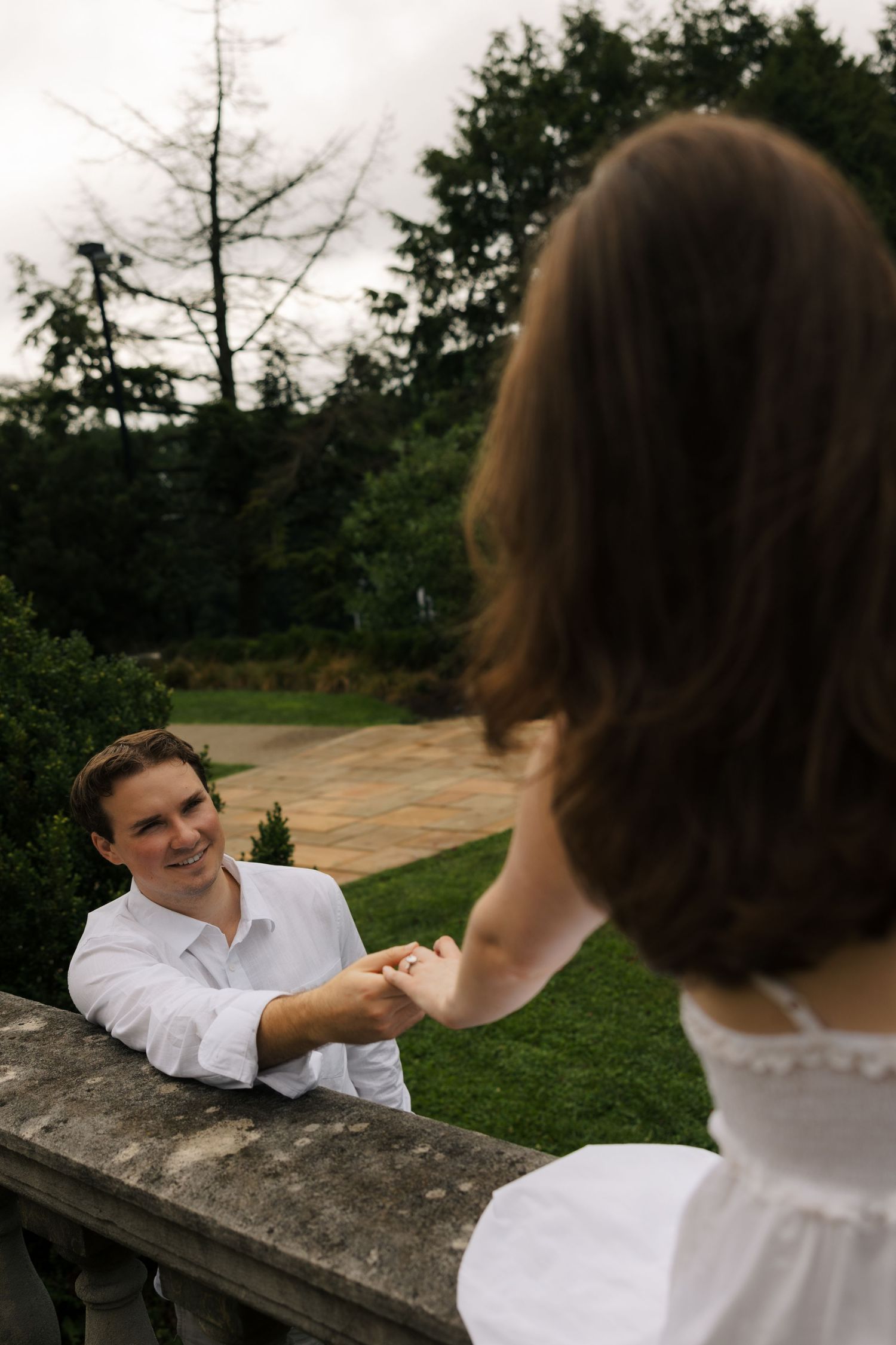Smiling man in white shirt holds womans hand over stone balustrade in lush garden setting.