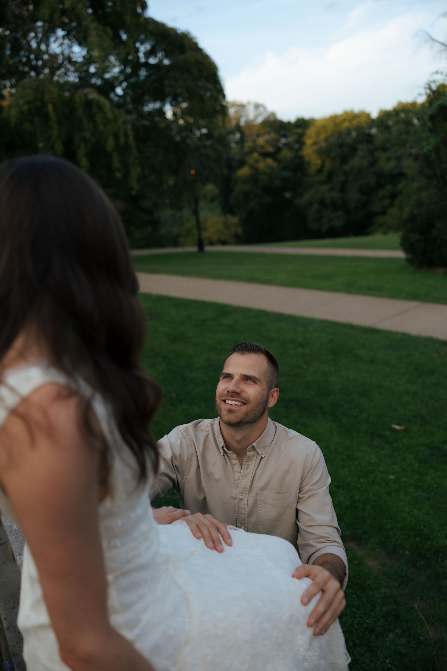 Smiling groom in beige shirt holding bride's white dress in lush green park at dusk.