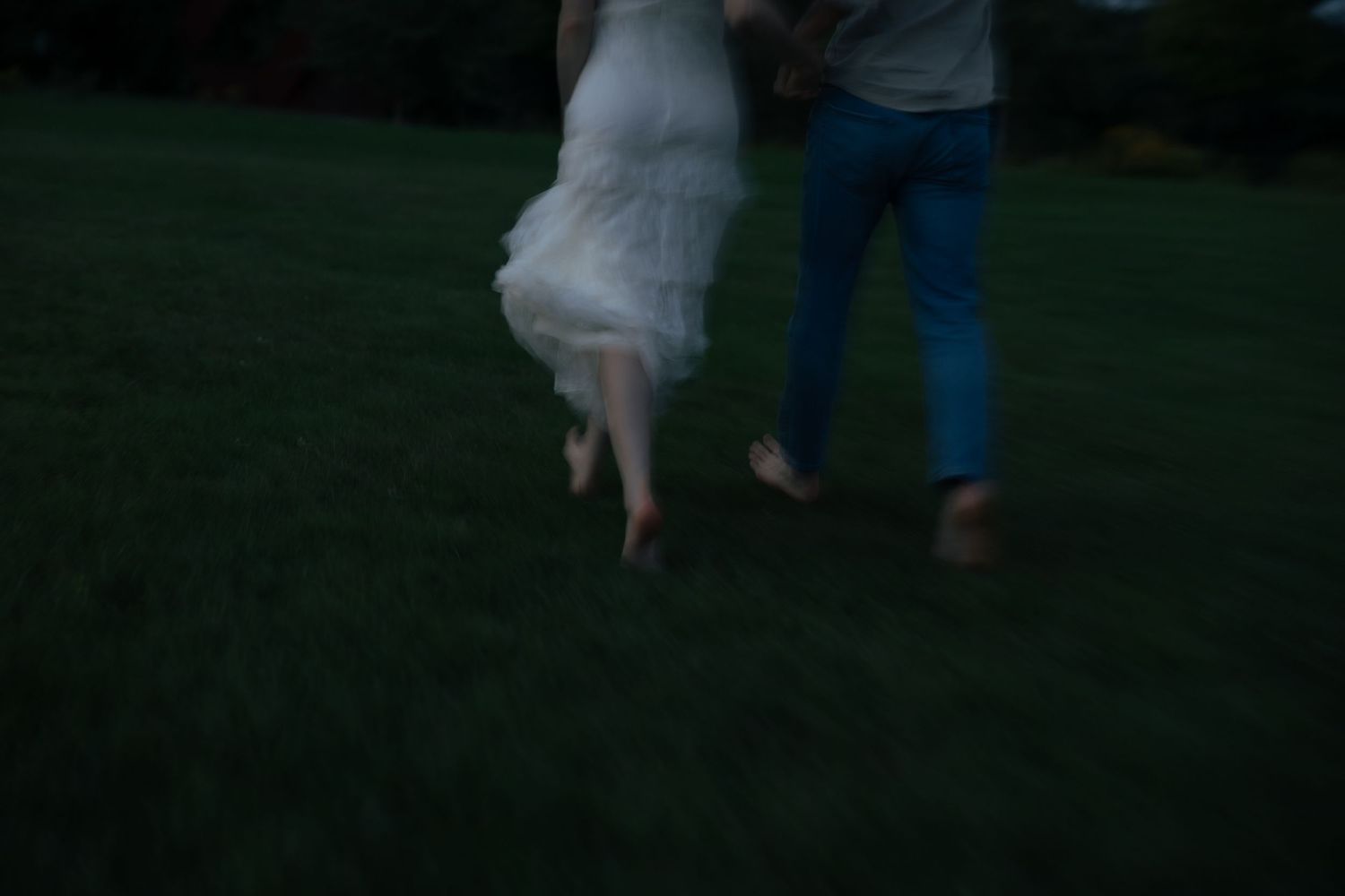 Blurred motion shot of barefoot couple running hand in hand across dark green lawn.