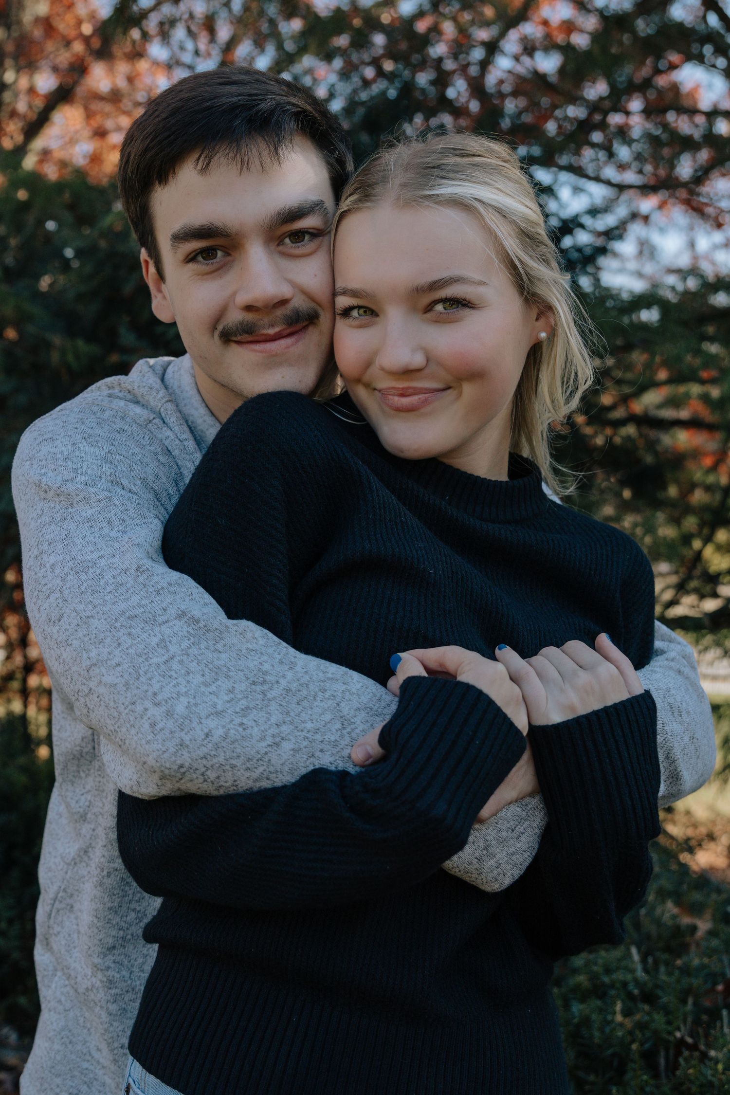 A couple embraces and shares tender moments outdoors during autumn with fall foliage in the background.