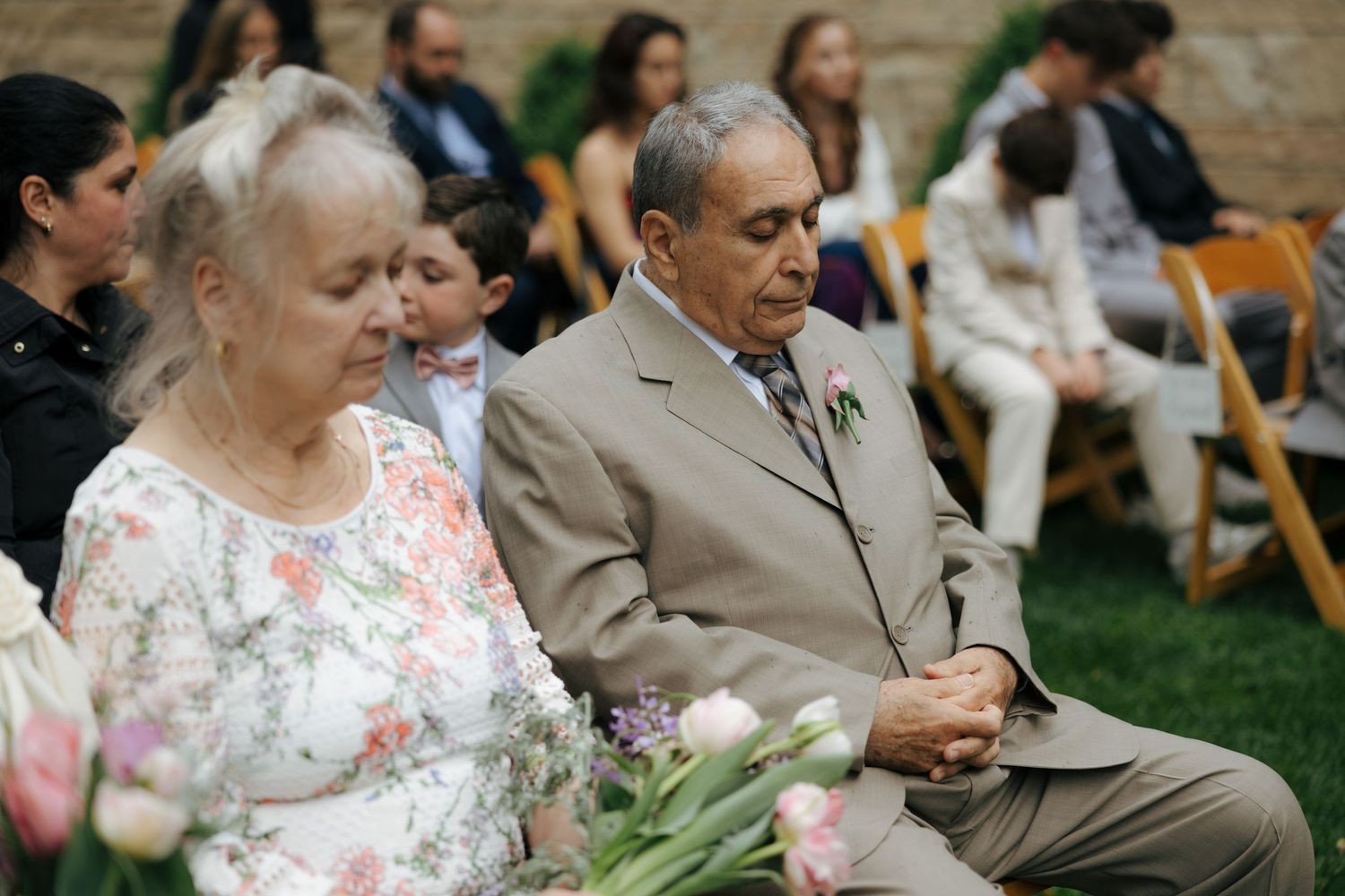 Elderly couple seated at outdoor wedding ceremony, man in tan suit holding tulips on his lap.