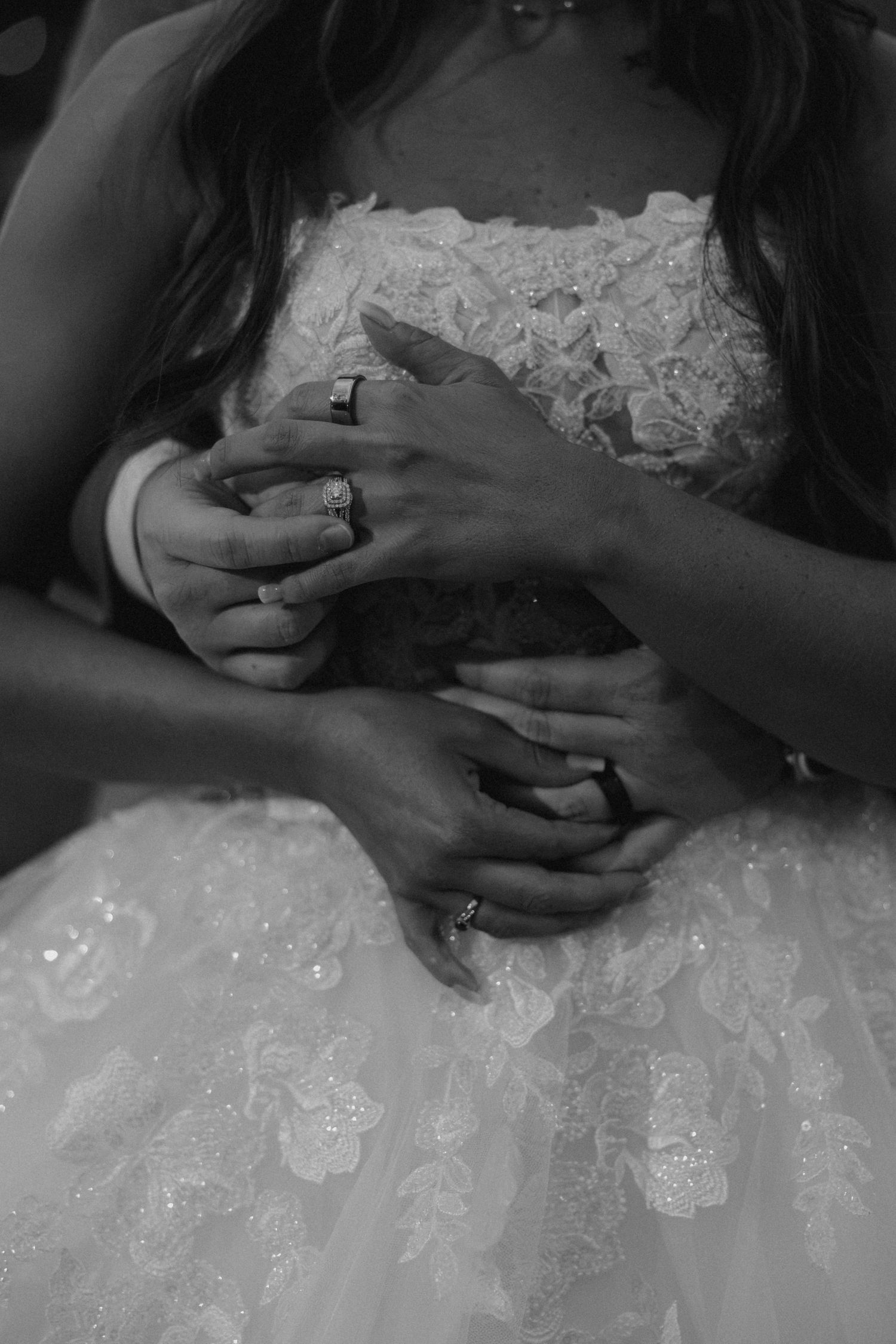 Black and white photo of bride and groom holding hands, showing wedding rings, with bride in lace gown.
