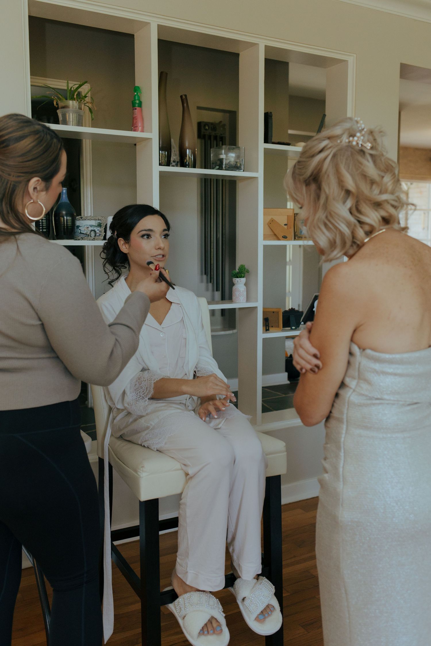 Bride getting makeup applied while bridesmaid in wedding dress stands nearby in bridal suite.