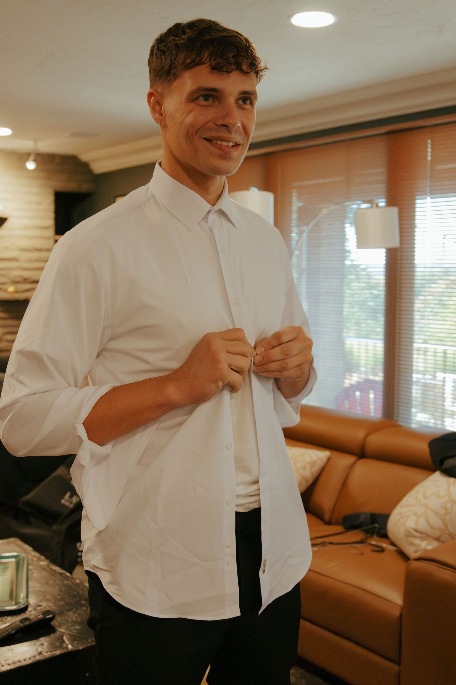 Smiling man buttoning white dress shirt in a living room, getting ready for a formal event.