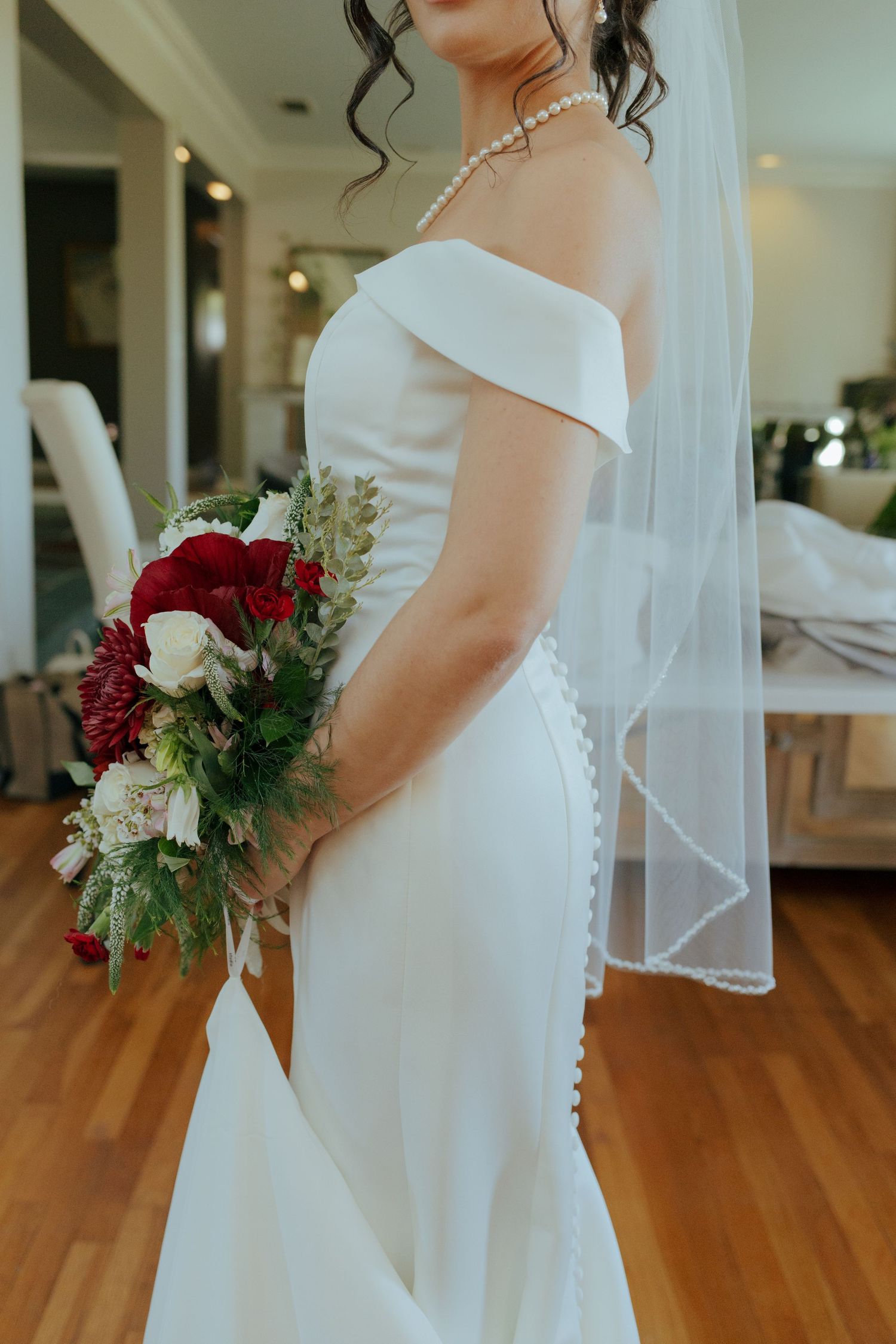 Bride in off-shoulder white mermaid gown holding red and white bouquet, wearing pearl necklace and veil.