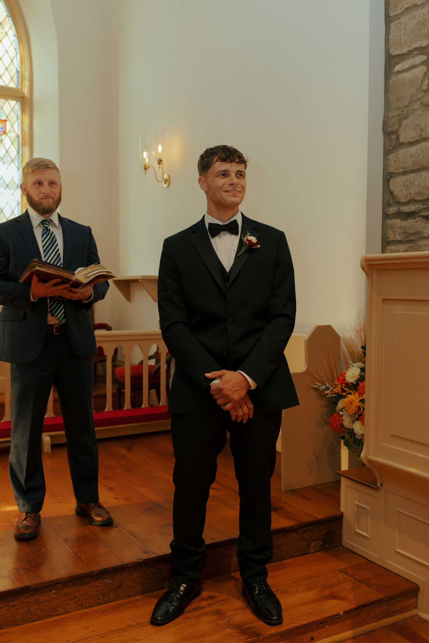Groom in black tuxedo smiling at altar in stone church, officiant holding book behind him.