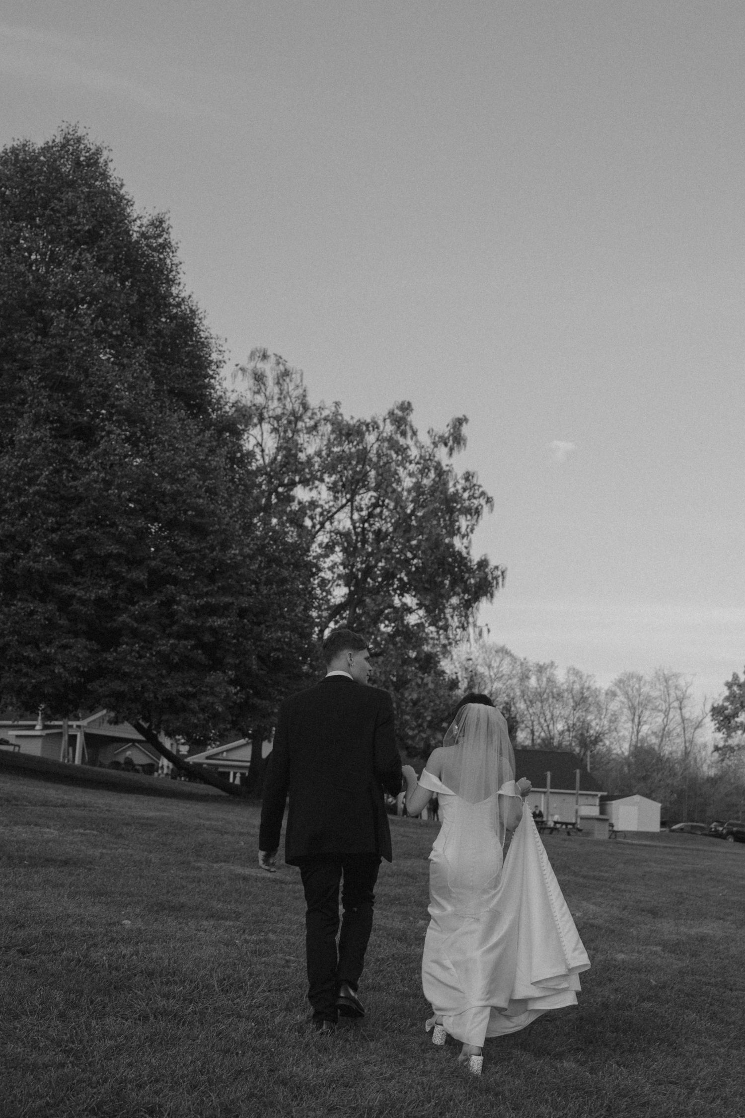 Black and white photo of bride and groom walking hand in hand across green lawn.