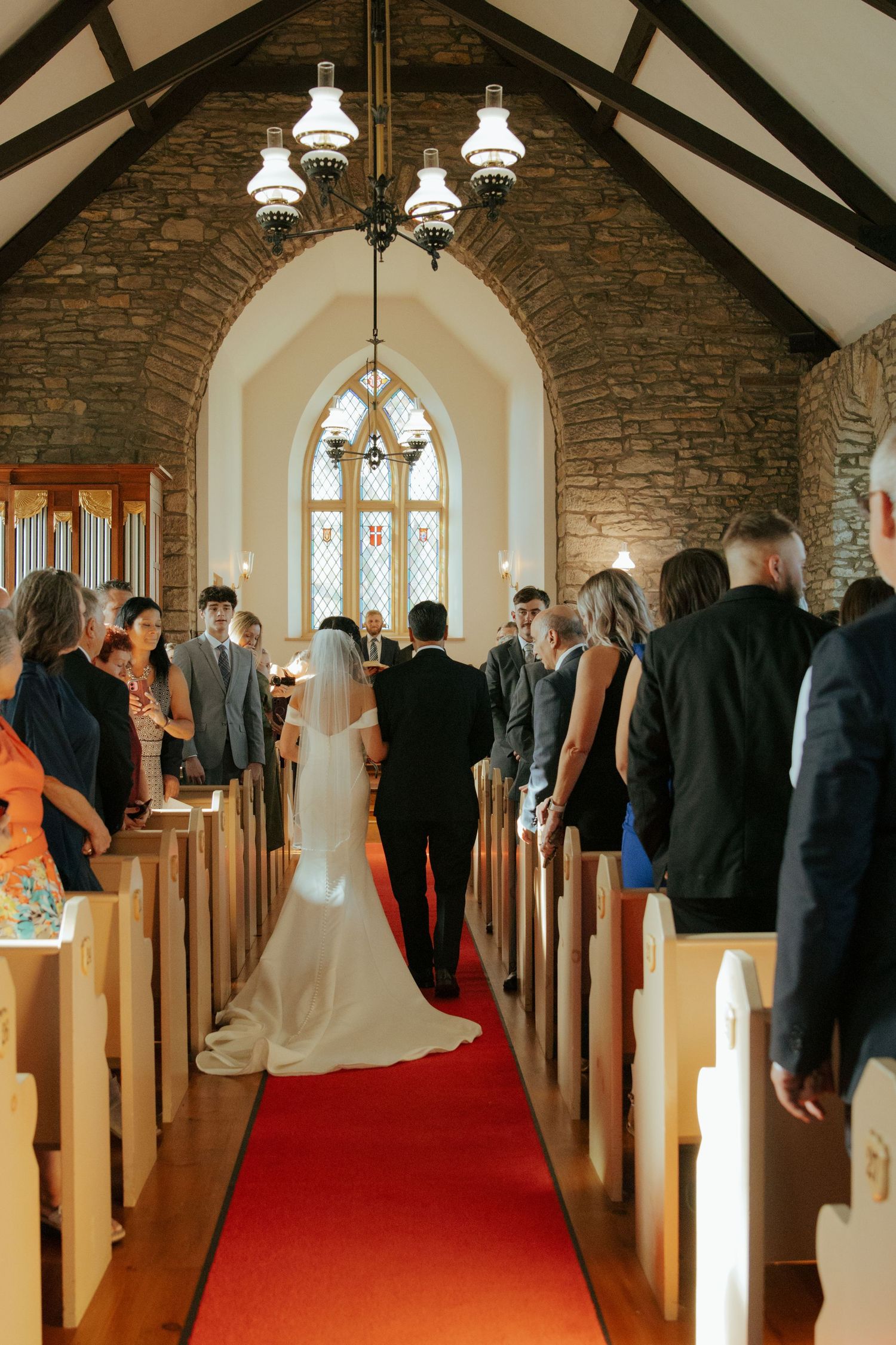 Bride walking down red carpet aisle of rustic stone chapel escorted during wedding ceremony.