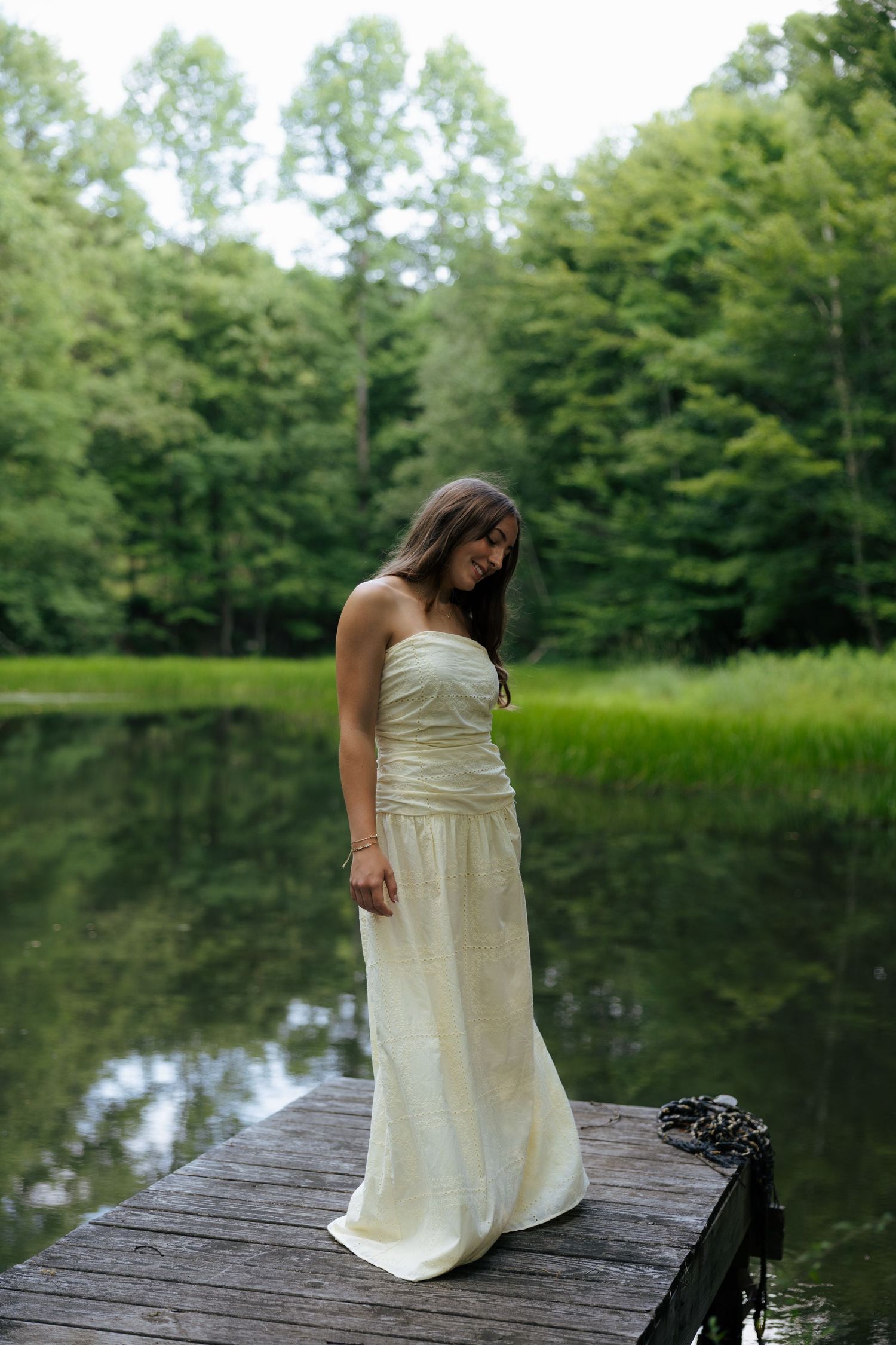 Young woman in elegant cream strapless gown standing on wooden dock overlooking calm forest lake.