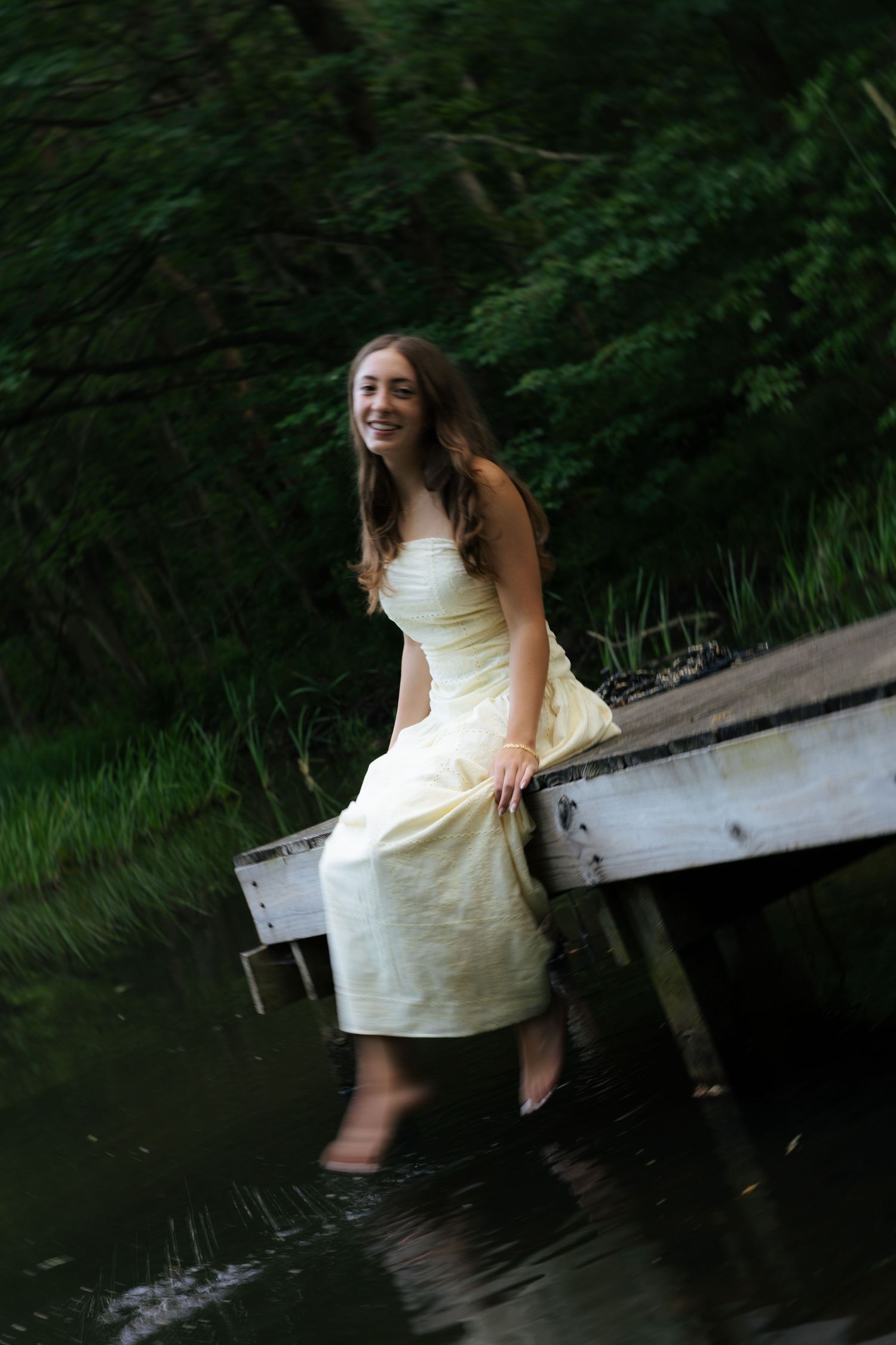 Laughing young woman in a yellow dress standing in shallow water beside an old wooden dock in a forest.
