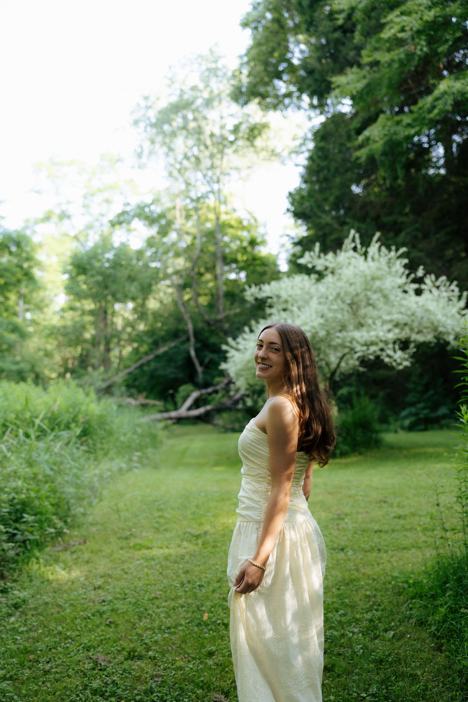 Smiling young woman in a white sundress standing on green grass surrounded by lush trees in a garden.