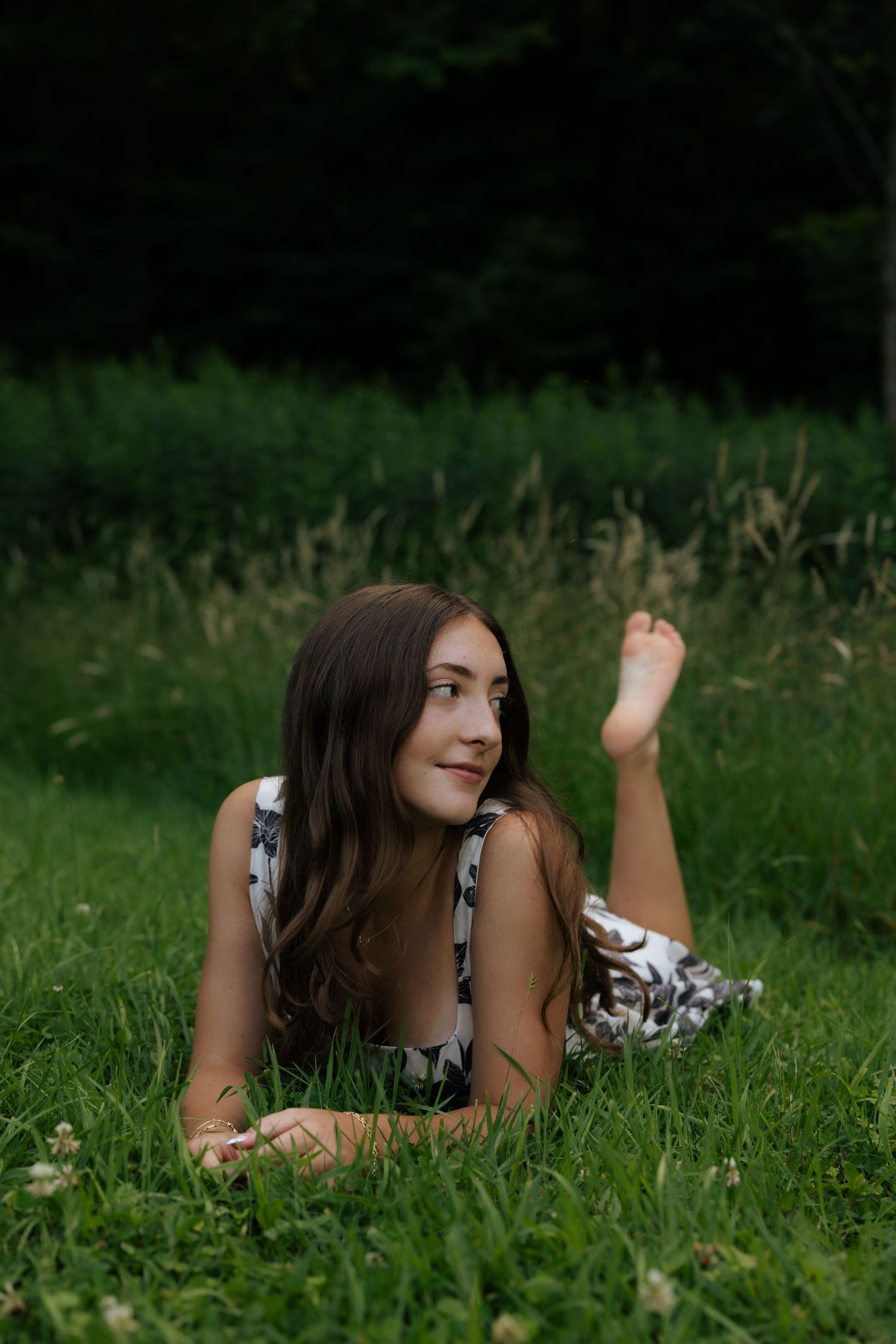 Young brunette woman lying on green grass in floral dress, looking sideways in a park setting.