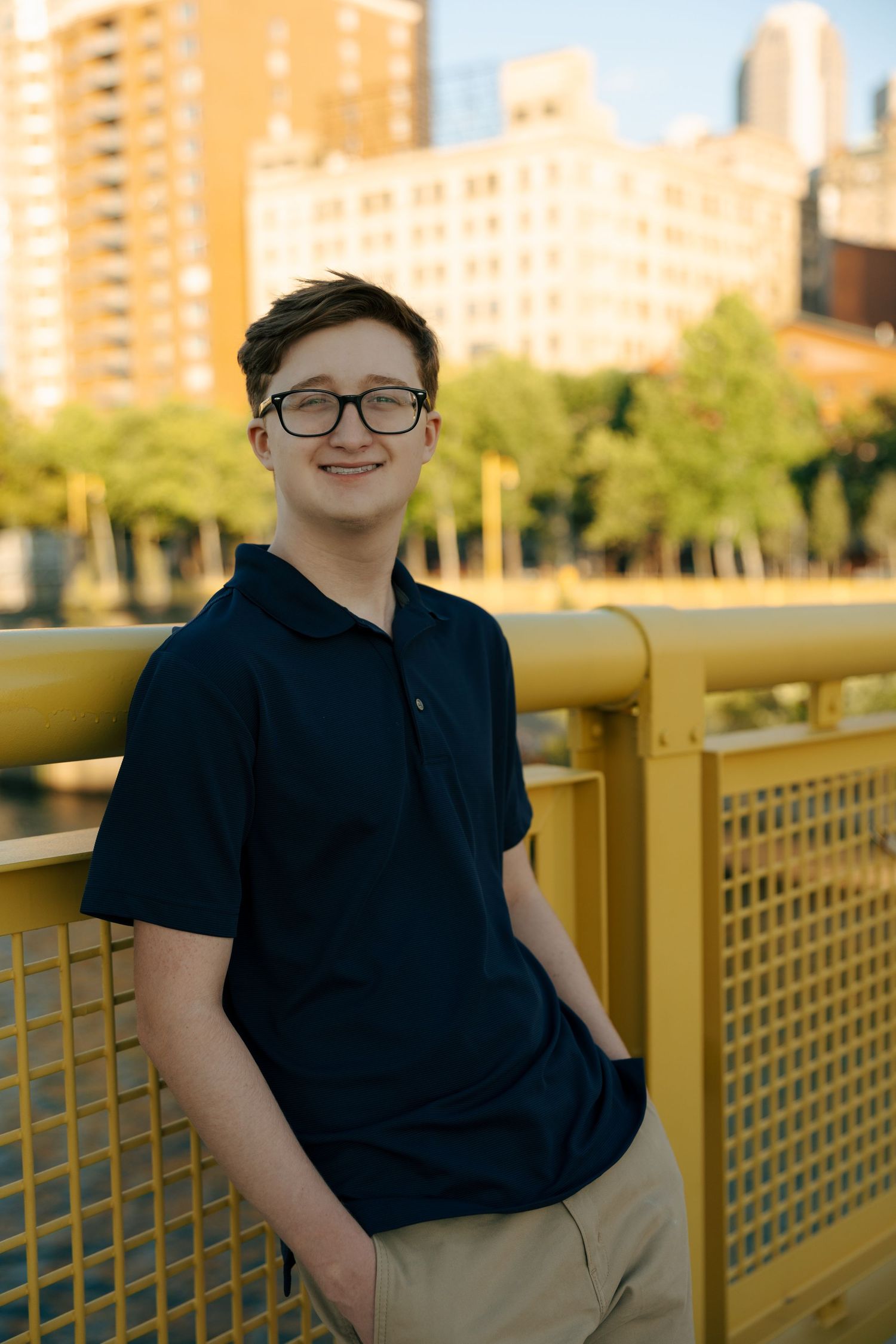 Smiling teen in navy polo and glasses leaning on yellow bridge railing with city buildings in background.