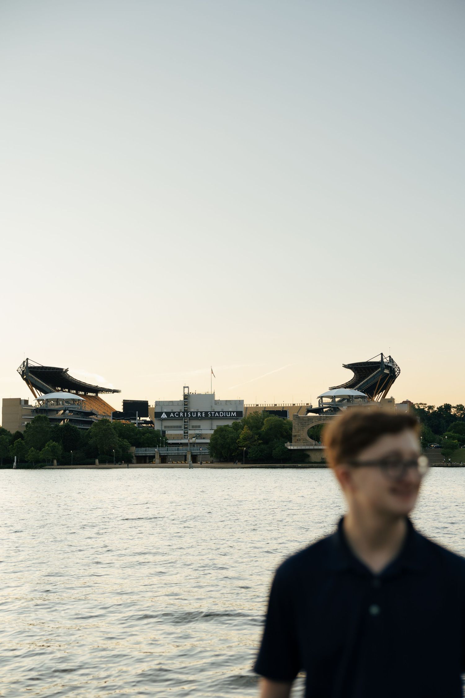 Acrisure Stadium across river at dusk with blurred teen in navy polo foreground.