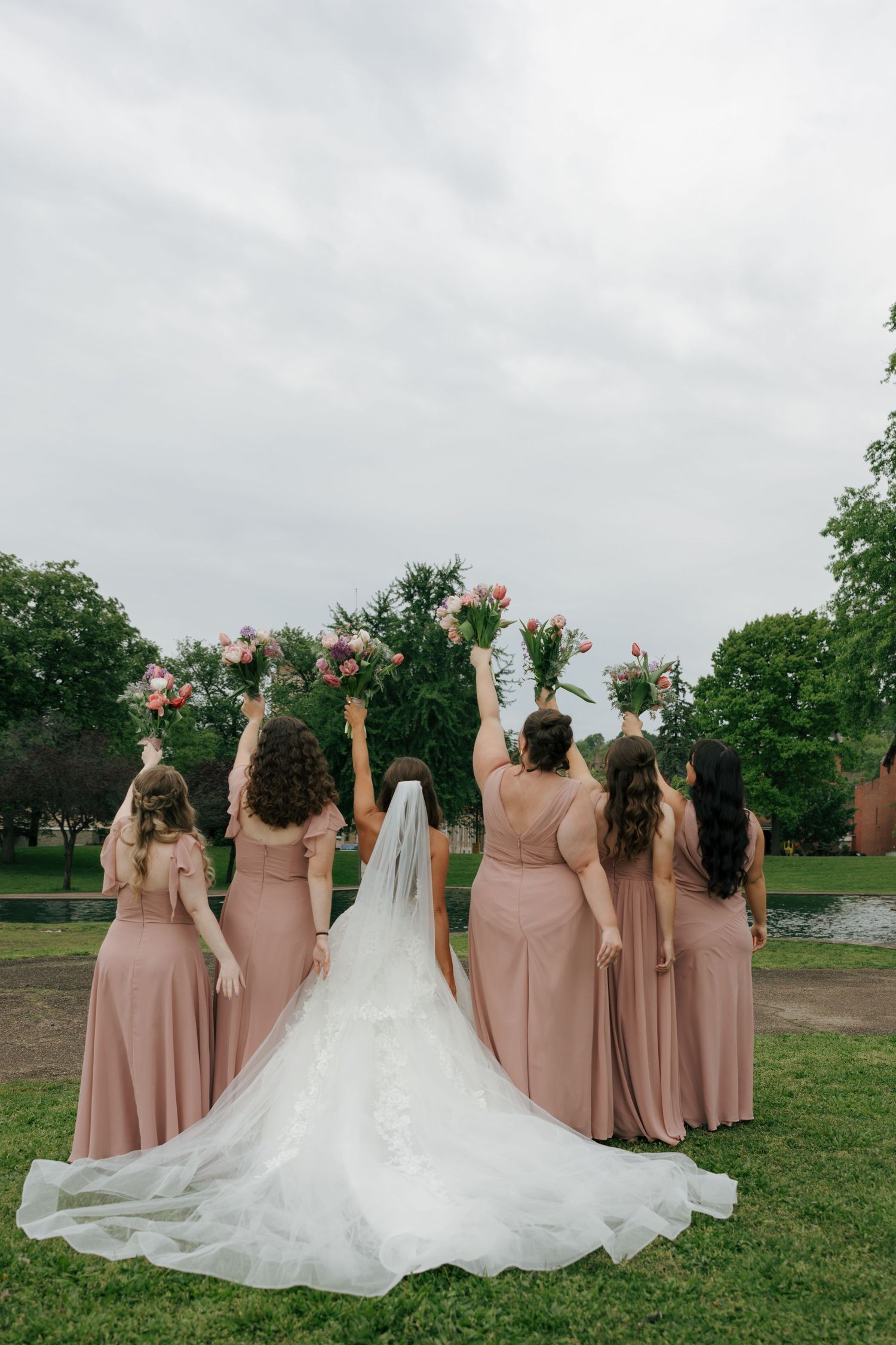 Bride and bridesmaids in blush gowns raise bouquets outdoors, viewed from behind.